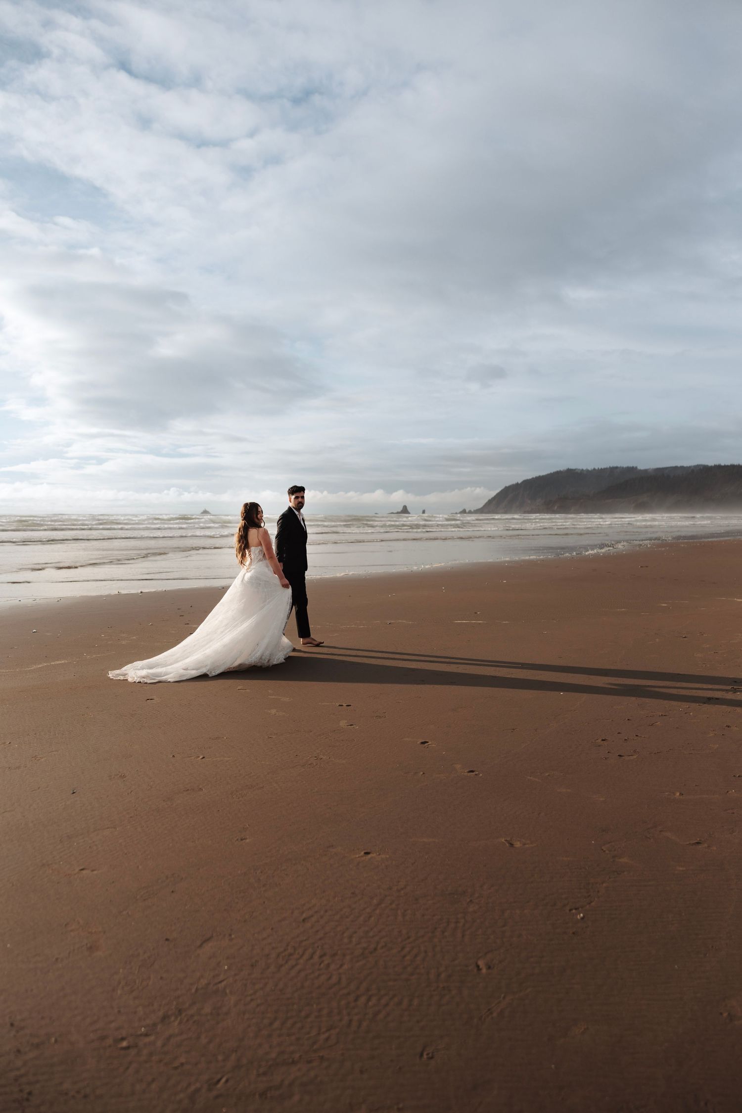Bride and groom in wedding attire walking hand in hand on vast sandy beach under cloudy sky.