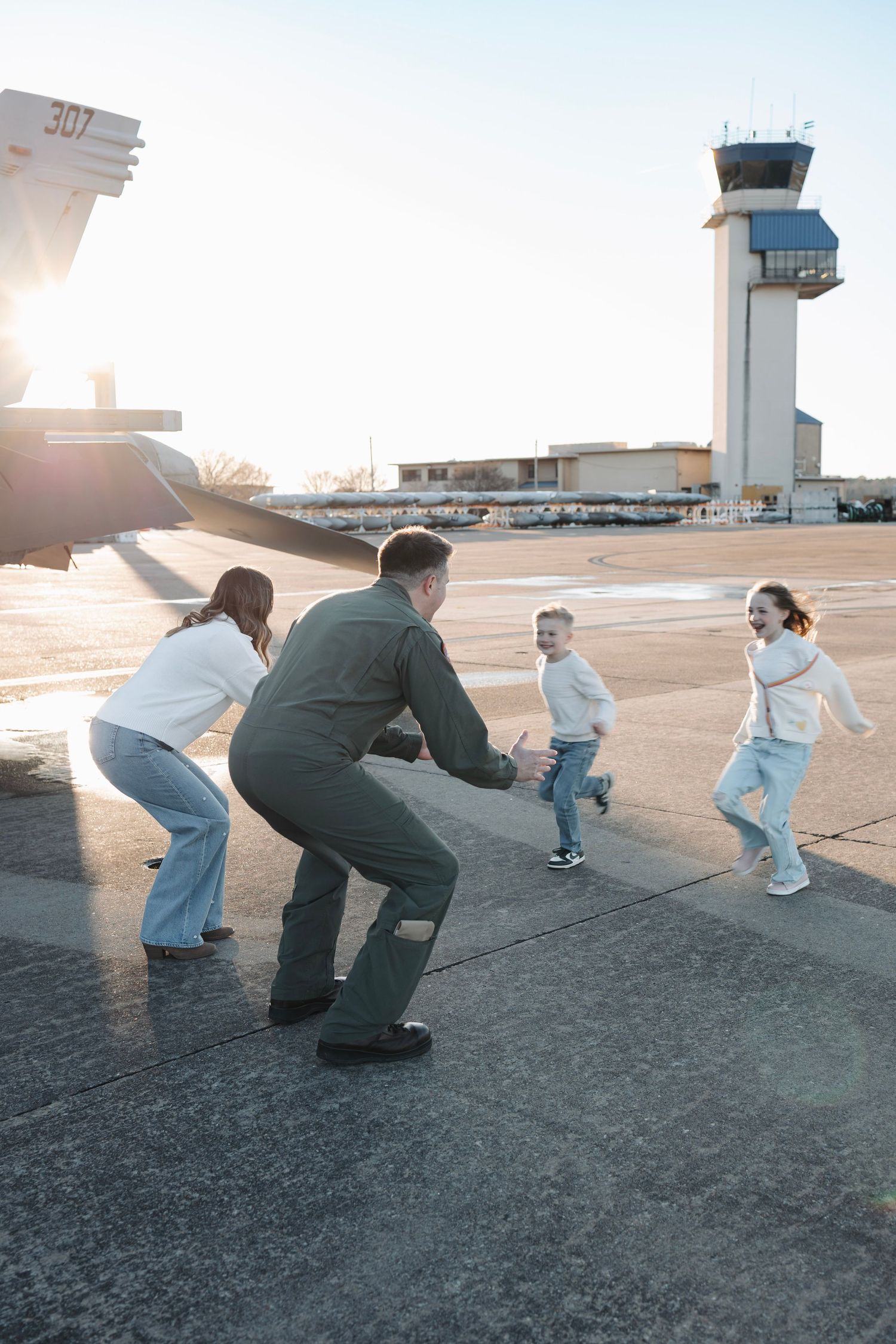 Military pilot in flight suit reuniting with family on airport tarmac at sunset.