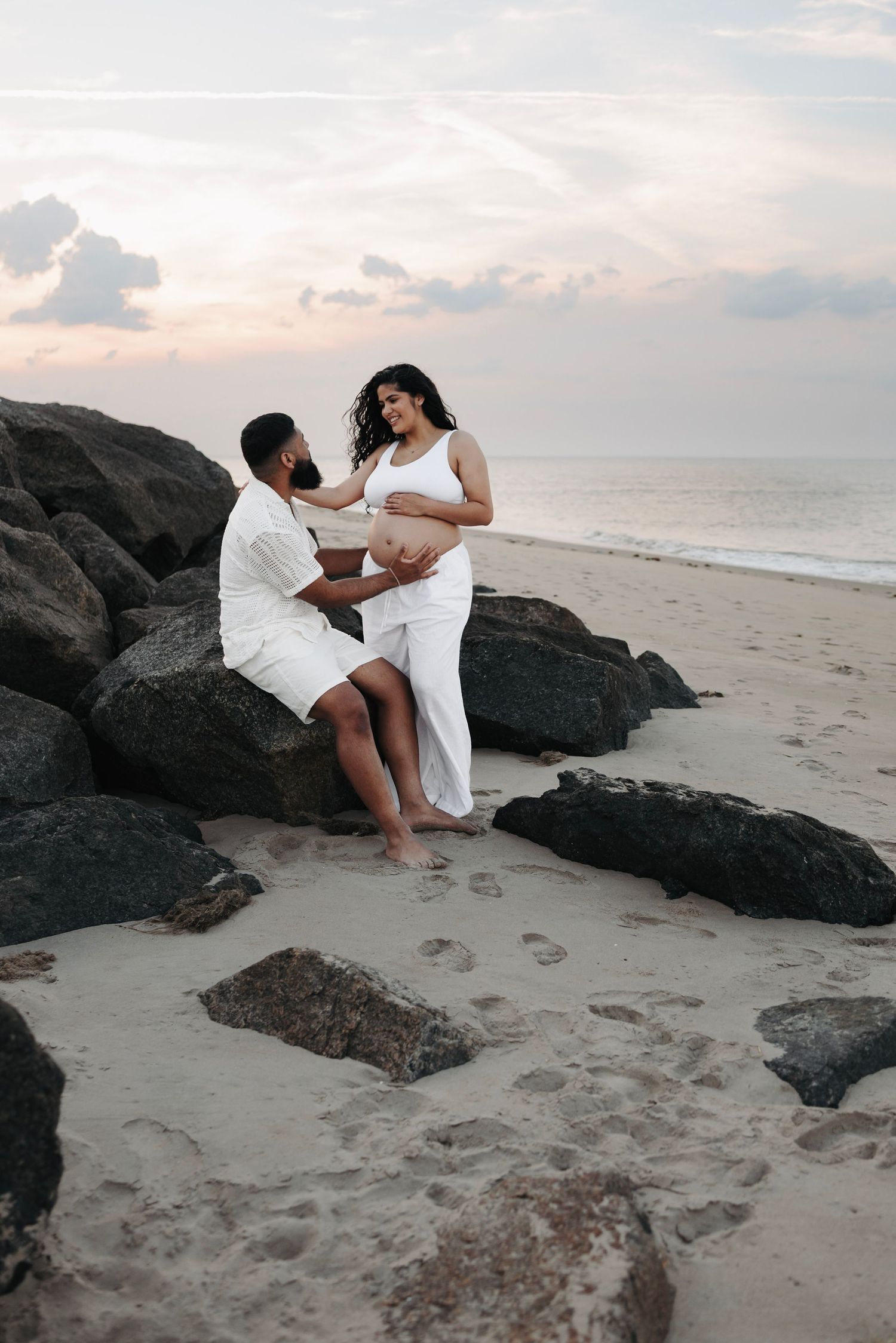 Expecting couple in white outfits sharing tender moment on rocky beach at sunset during maternity shoot.