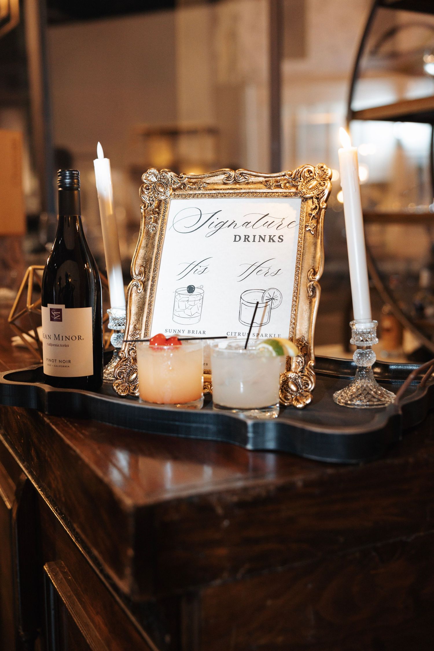Elegant wedding signature drinks display in gold frame with candles and cocktails.