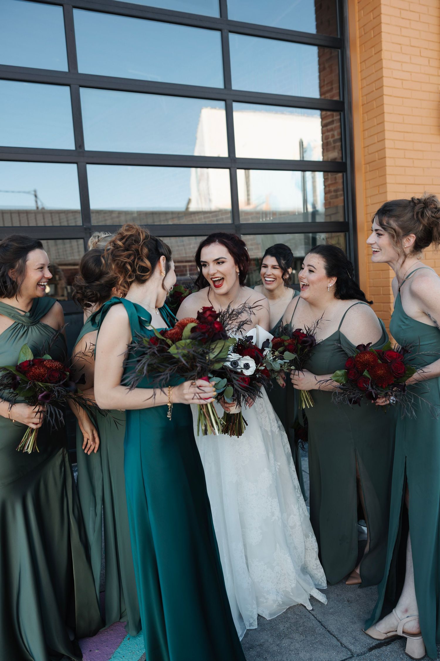 Bride in white gown laughing with bridesmaids in green dresses holding red bouquets outside brick building.