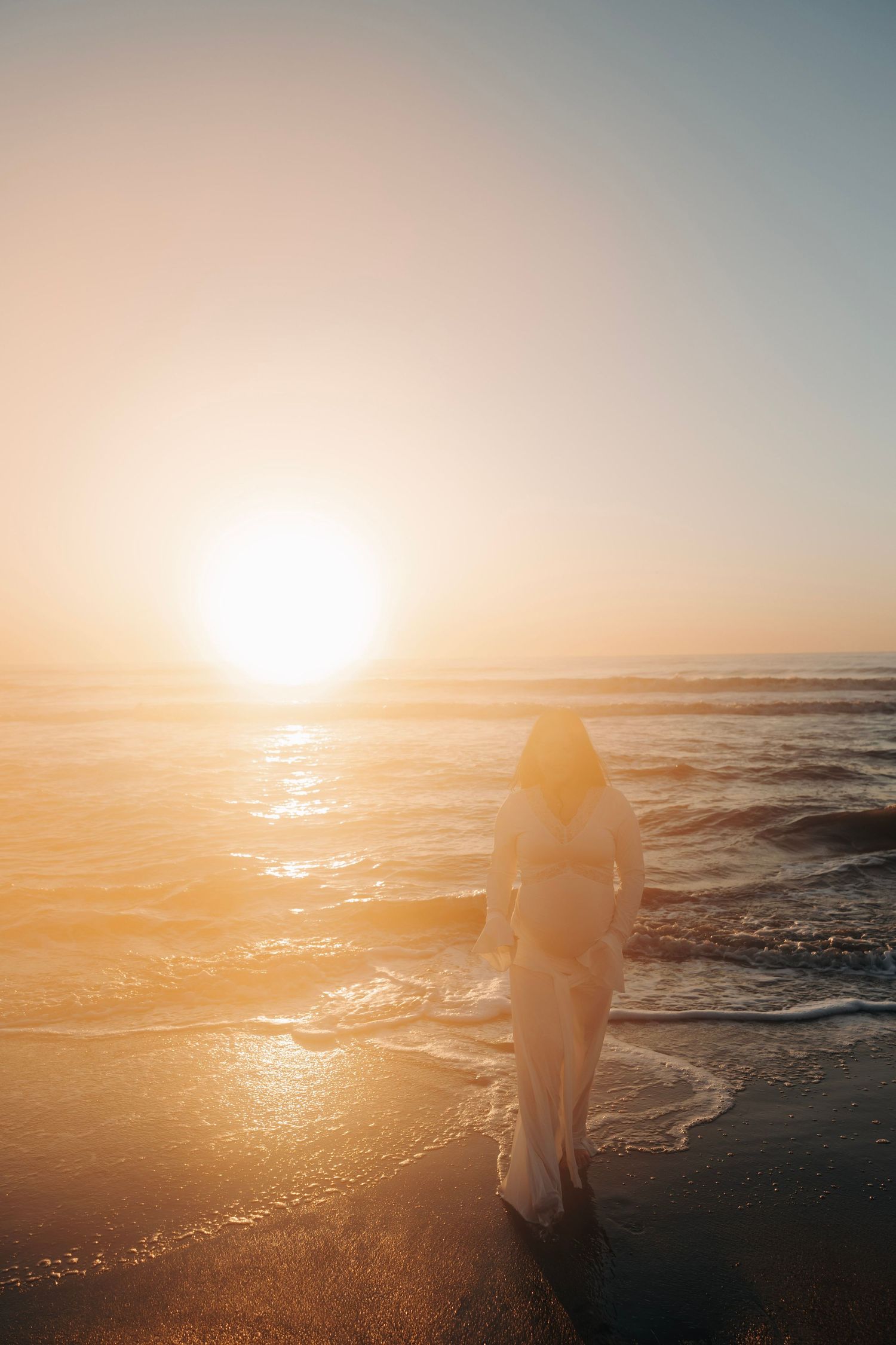 Woman in white dress walking toward golden sunset waves on a serene beach.