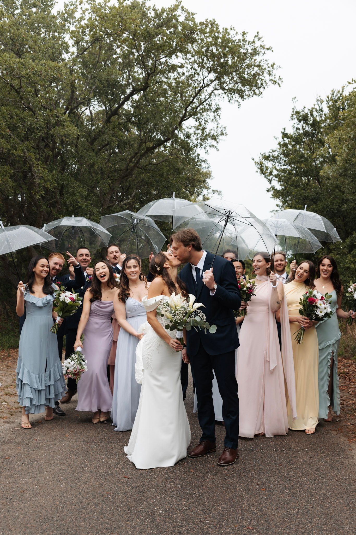 Wedding party kissing in rain with clear umbrellas, bridesmaids in pastel gowns on tree-lined path.