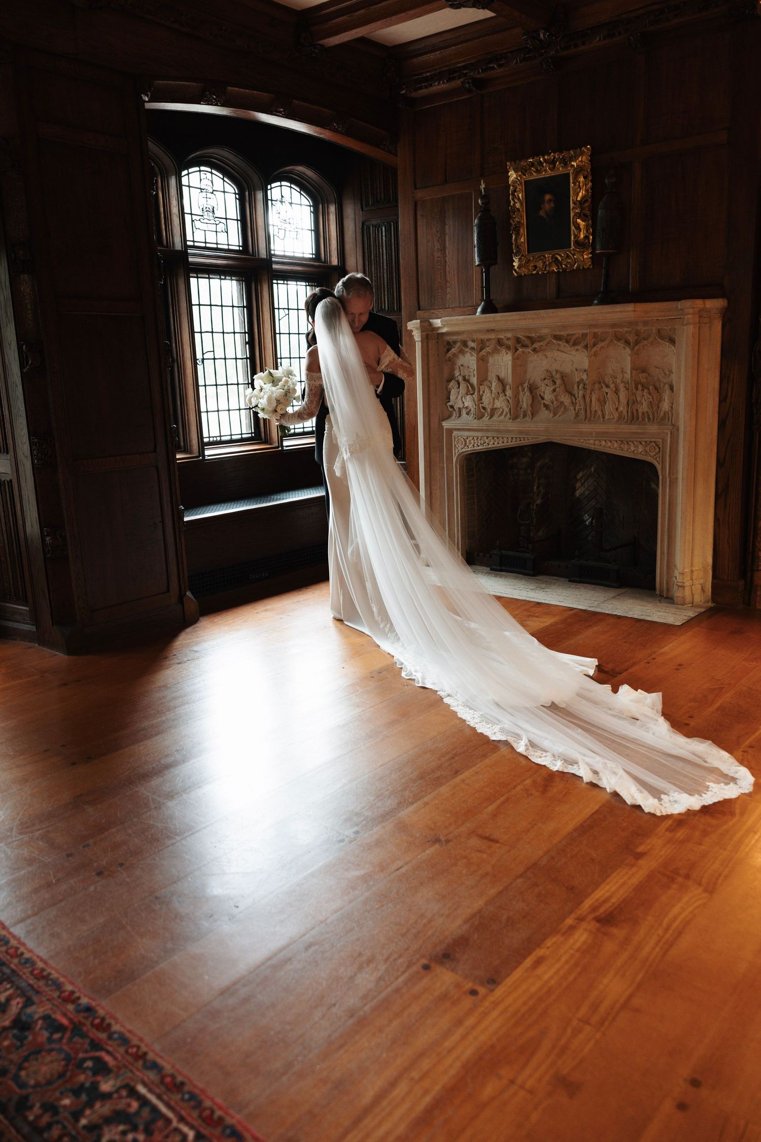 Bride in flowing white gown and long veil stands near ornate fireplace in elegant wood-paneled room.