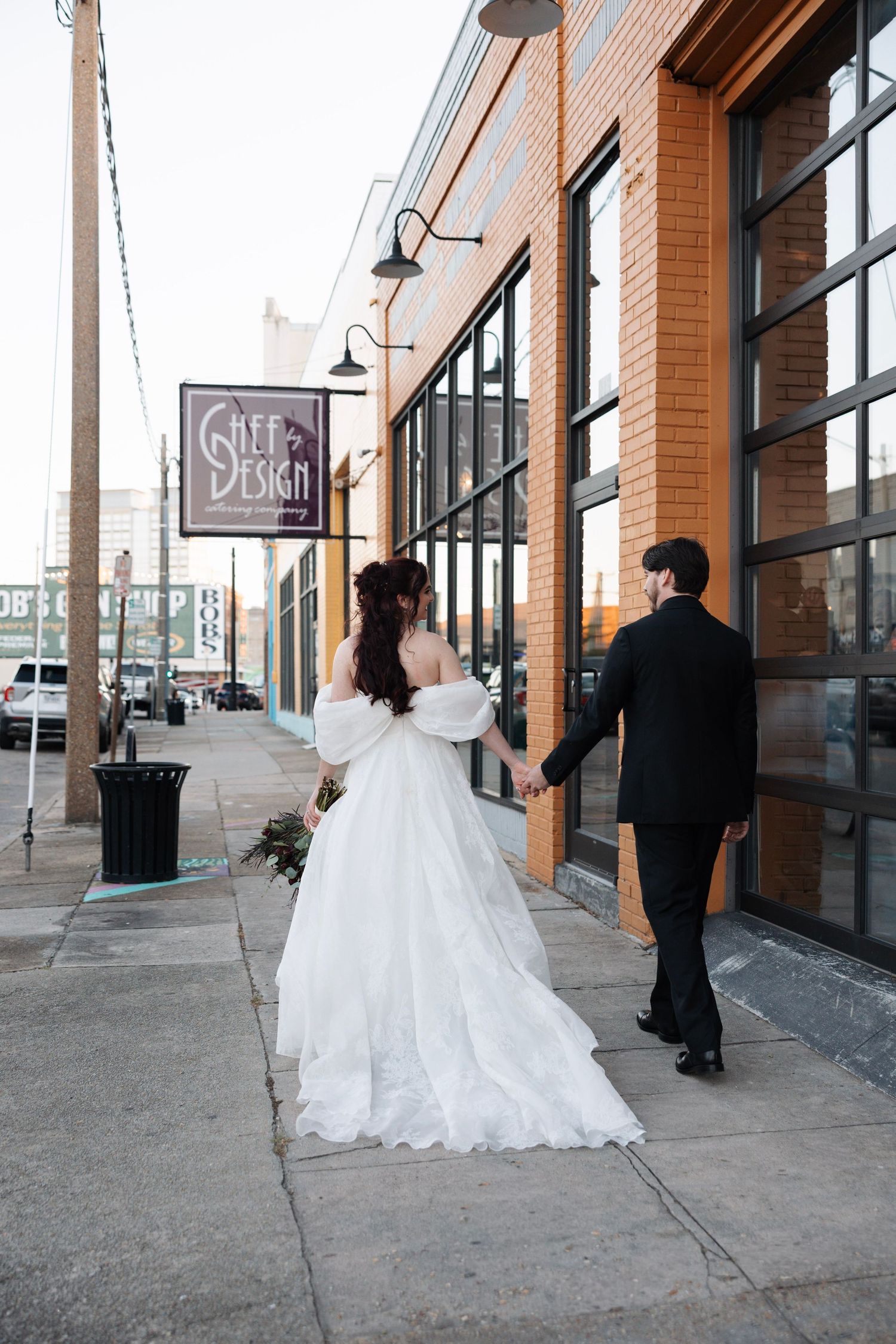 Bride and groom holding hands walking along urban sidewalk past brick building at sunset.