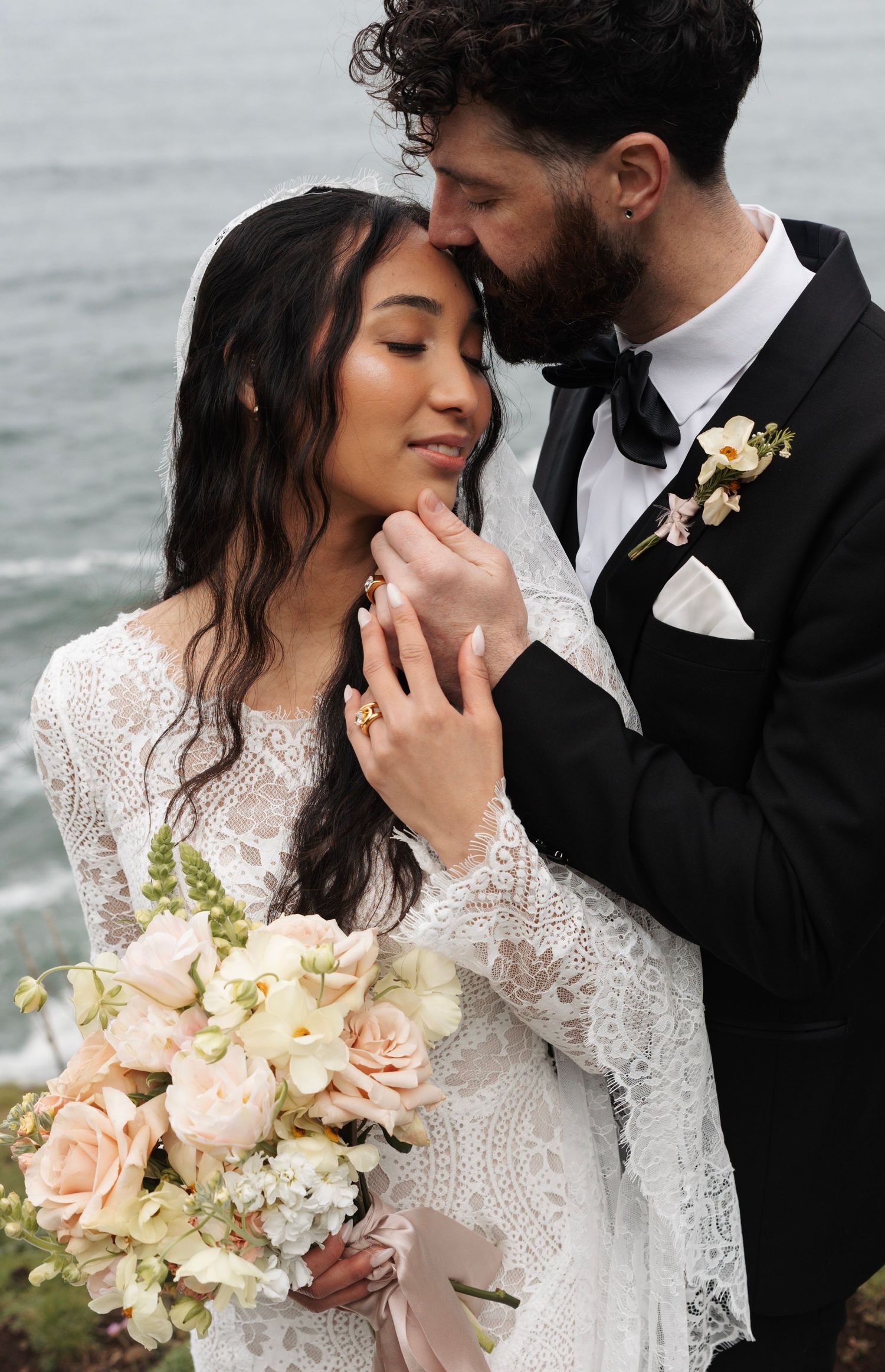 Groom in black tuxedo tenderly kissing bride holding blush bouquet by ocean.