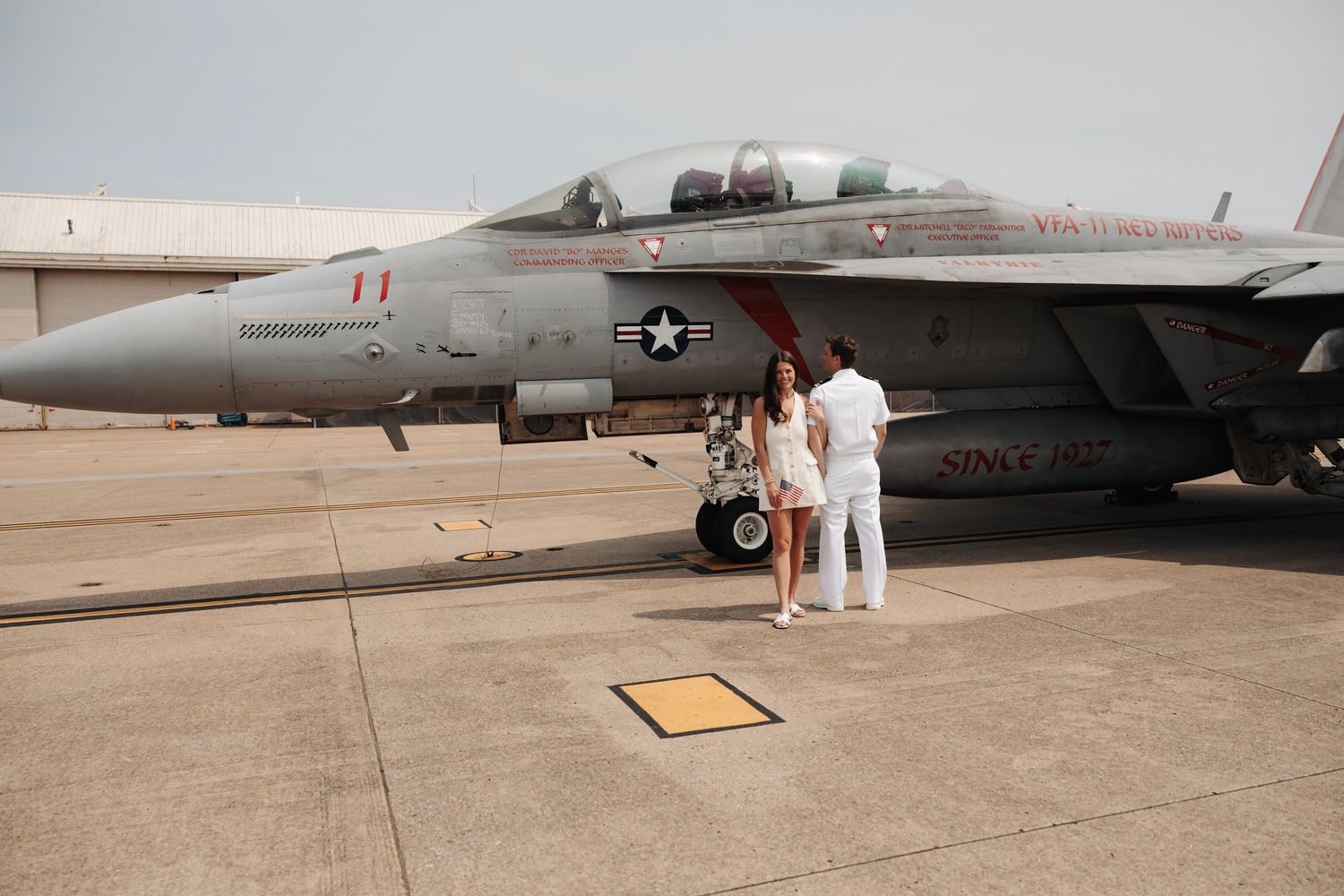 Couple standing before a US Navy F/A-18 fighter jet on an airfield tarmac.