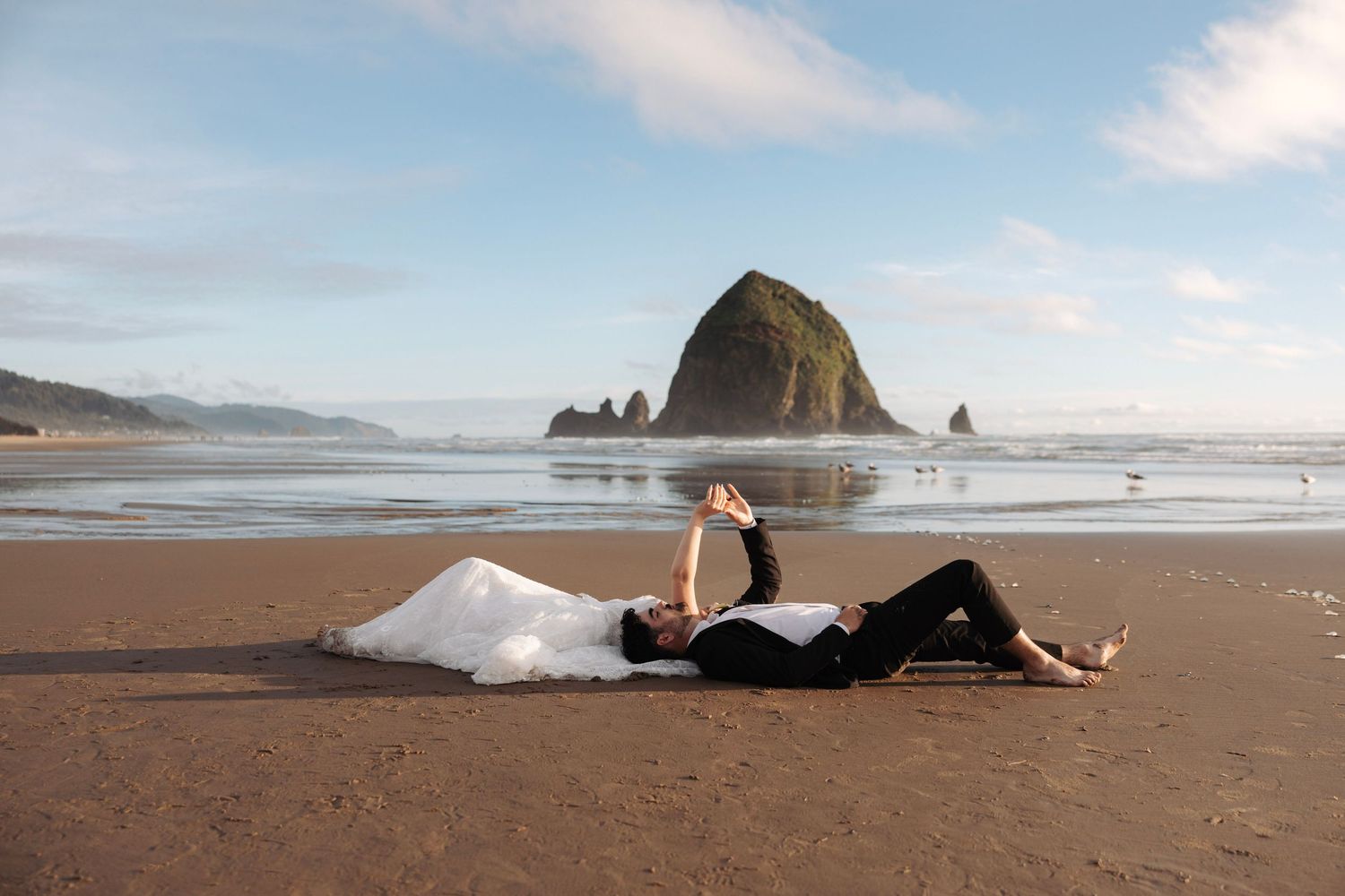 Bride and groom lying on sandy beach with Haystack Rock and blue sky in background.