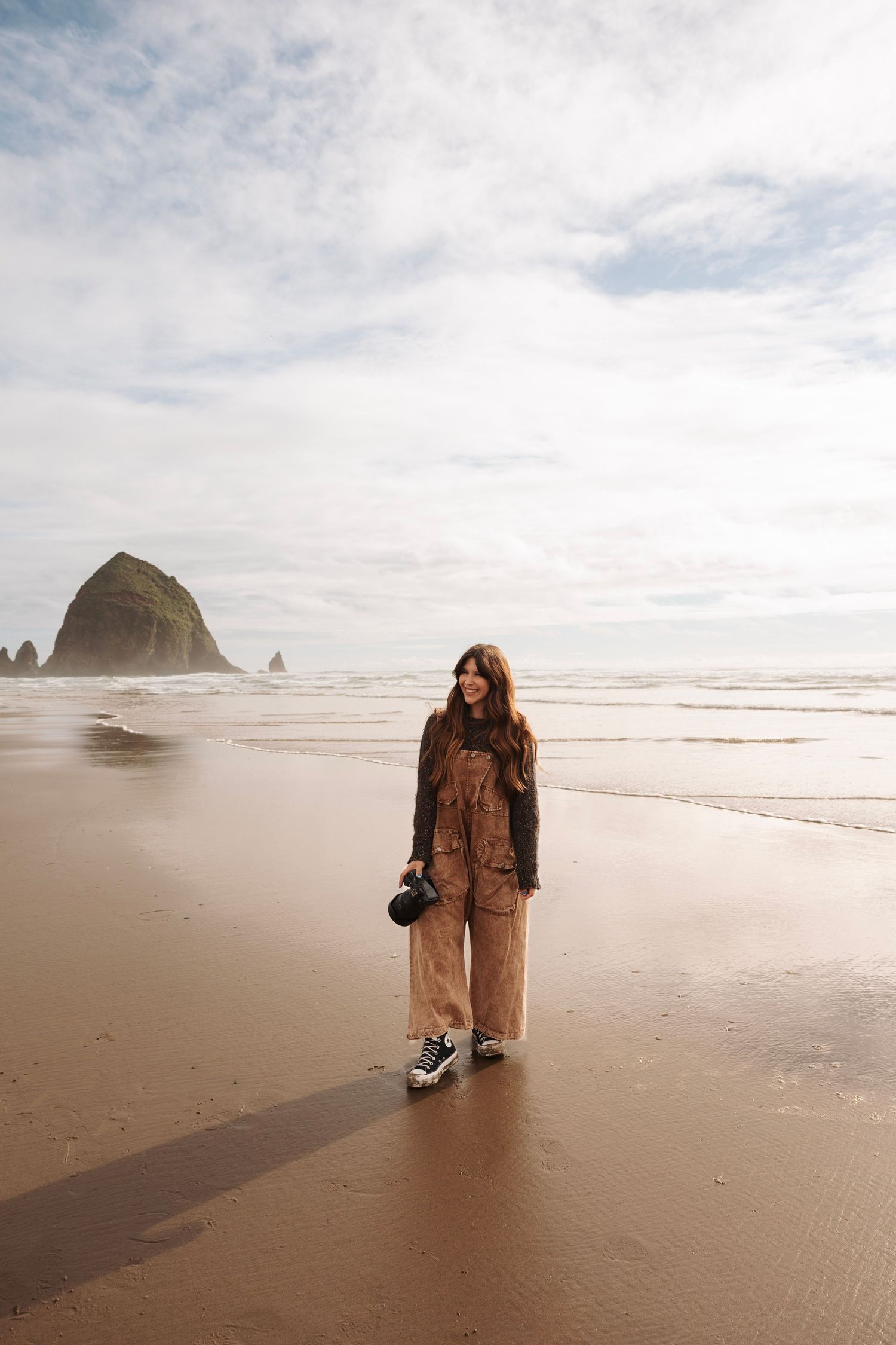 Smiling woman in tan overalls holding camera on sandy beach with iconic Haystack Rock behind her.