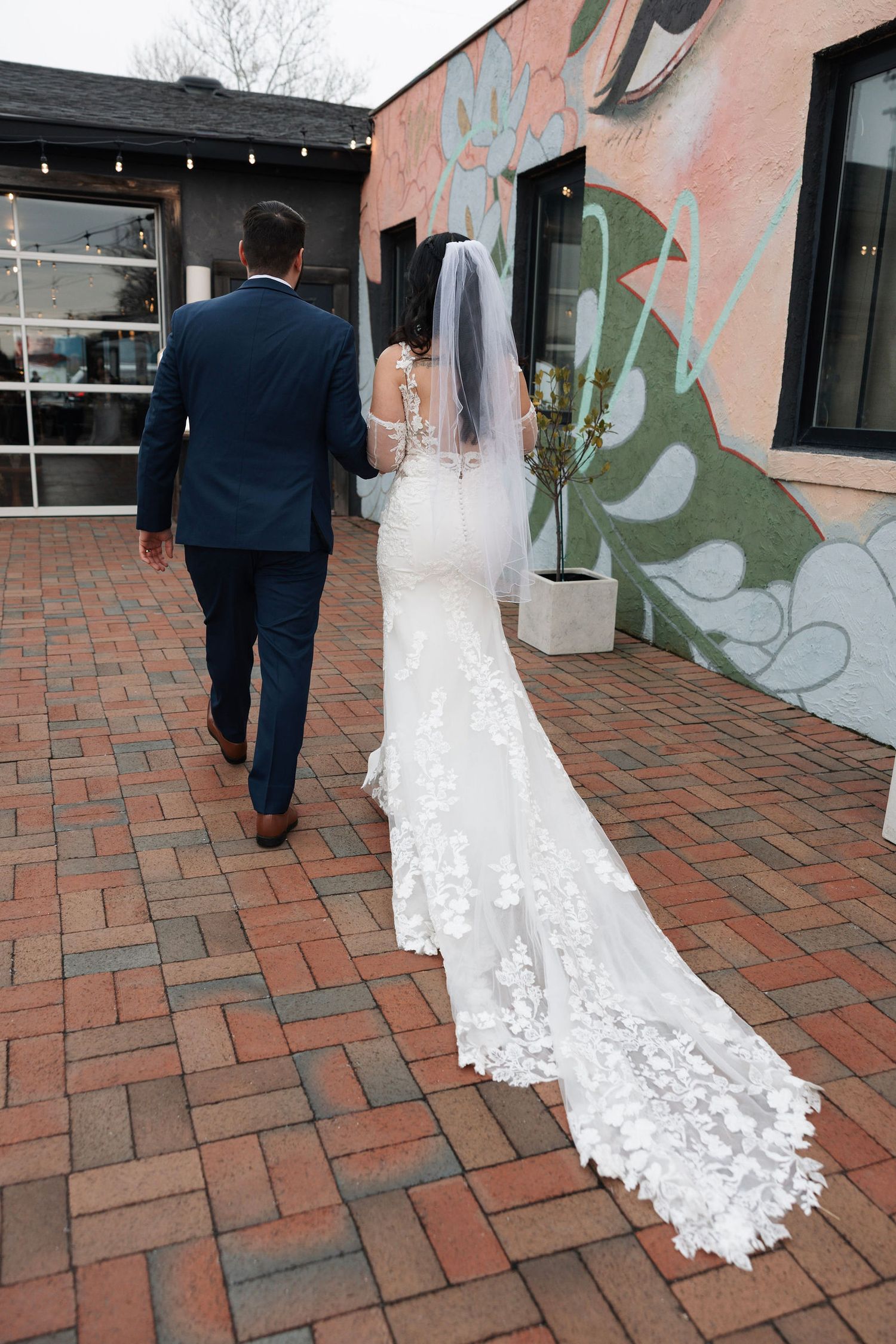 Bride in lace gown with cathedral veil walking with groom past colorful mural on brick patio.