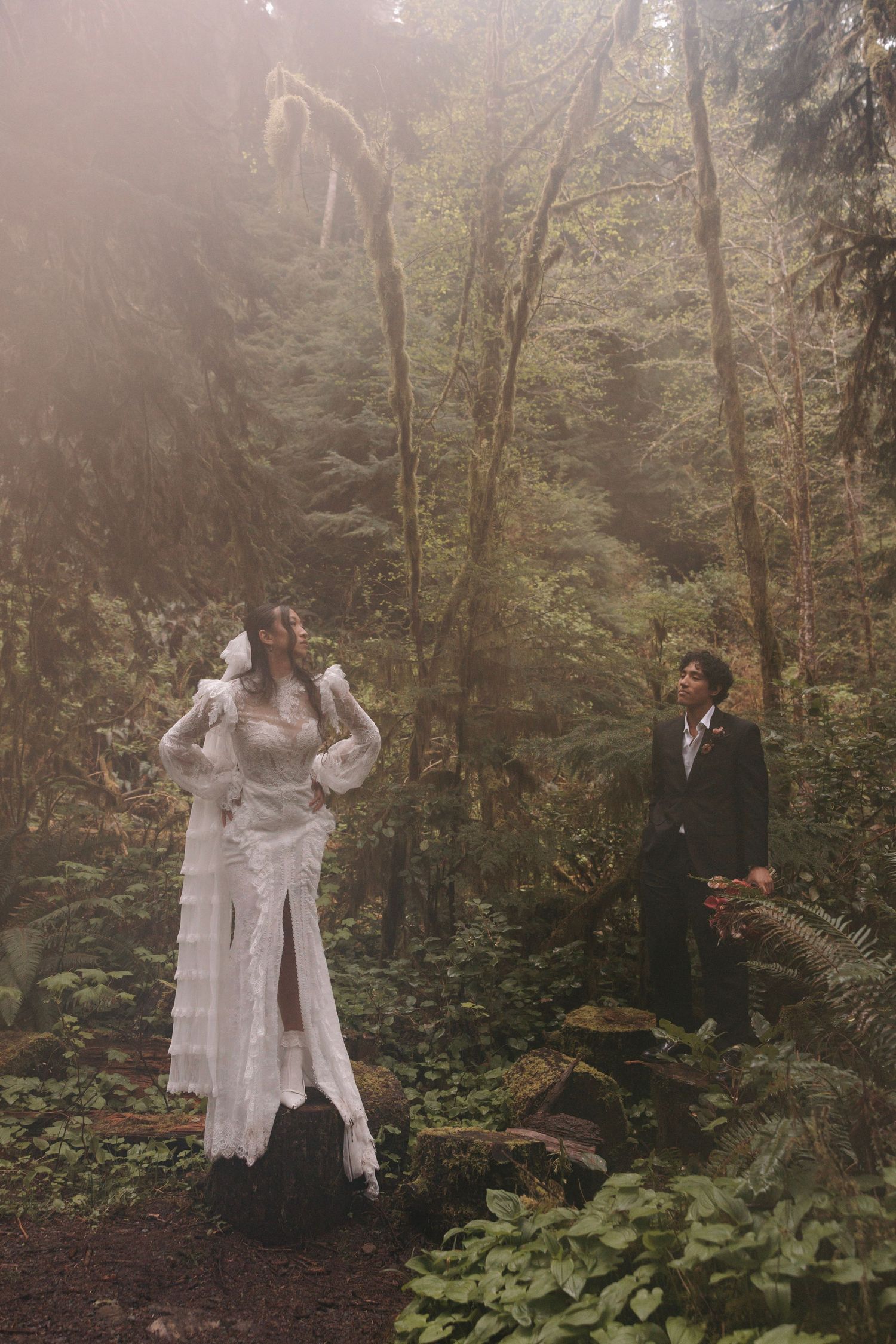 Bride and groom stand among lush forest trees in ethereal misty light, bride wearing elegant lace wedding gown.