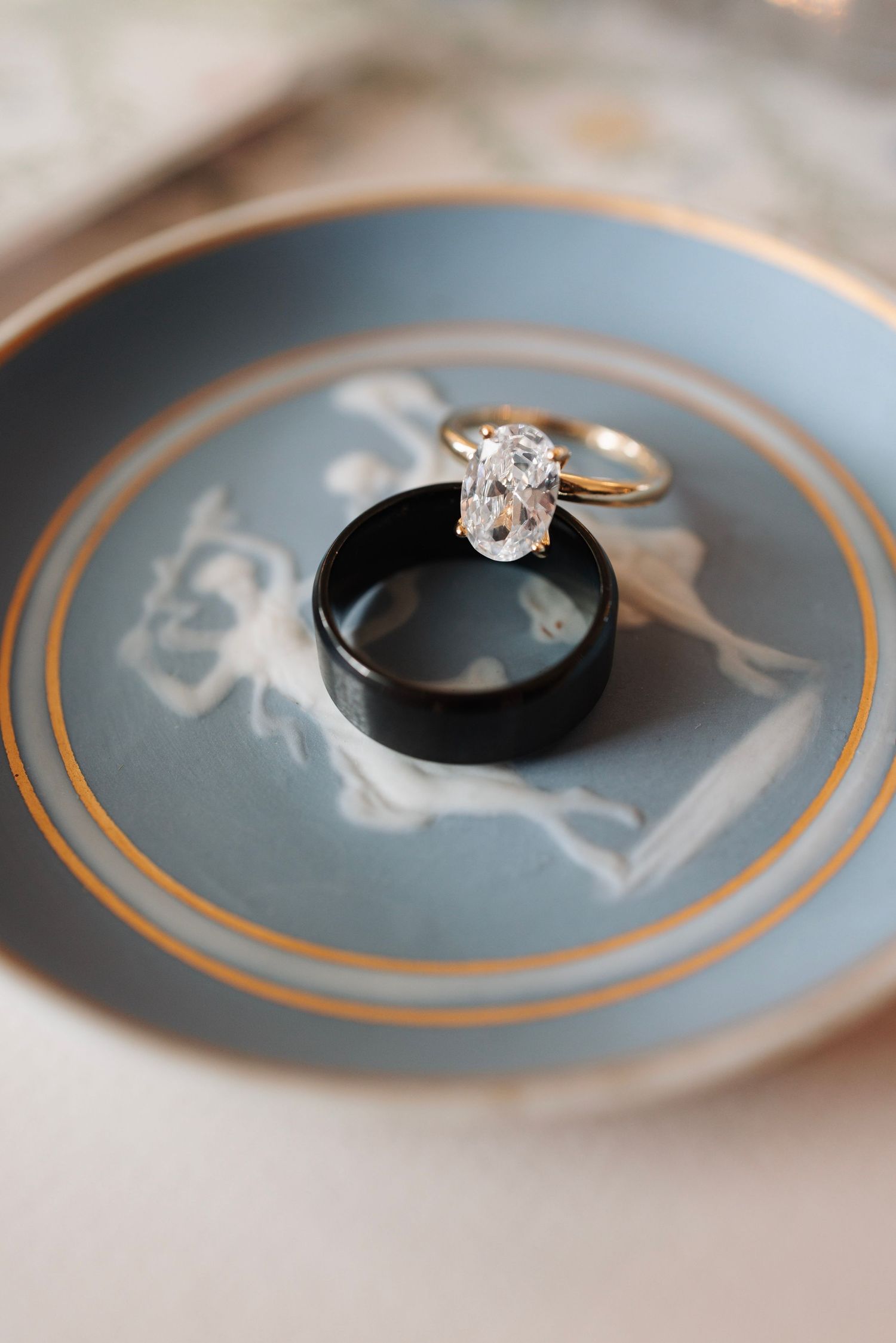 Gold diamond engagement ring and black wedding band displayed on a vintage blue decorative plate.