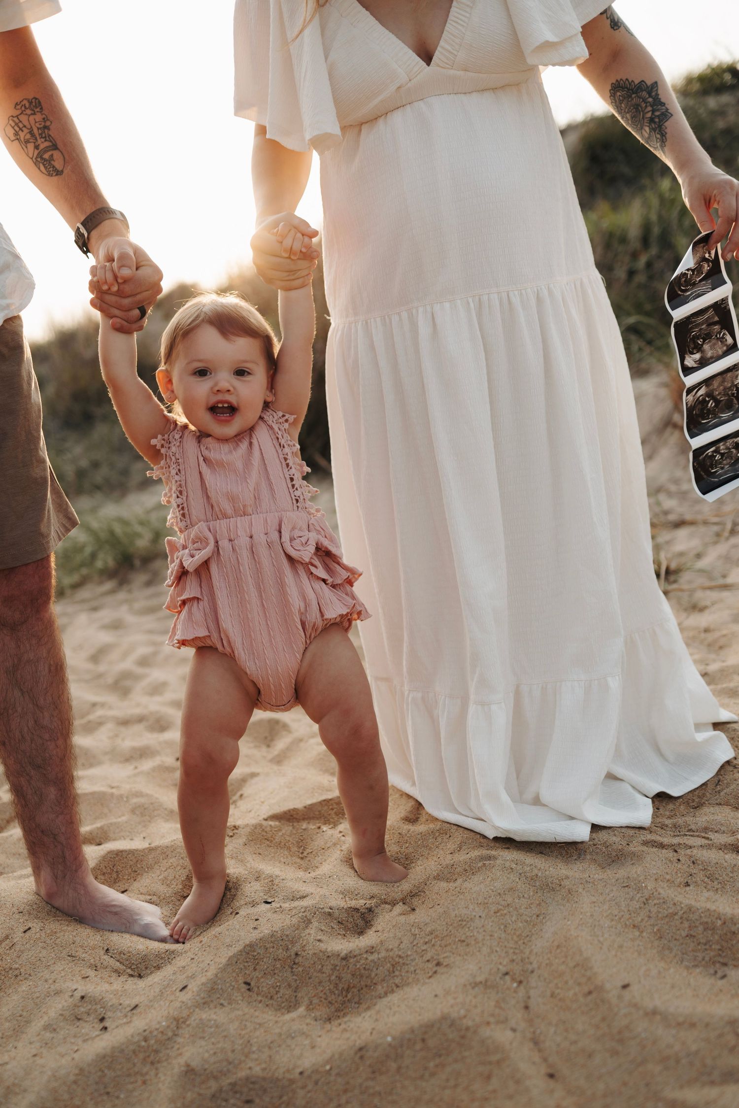 Happy baby girl in pink romper taking first steps on sandy beach held by parents at sunset.