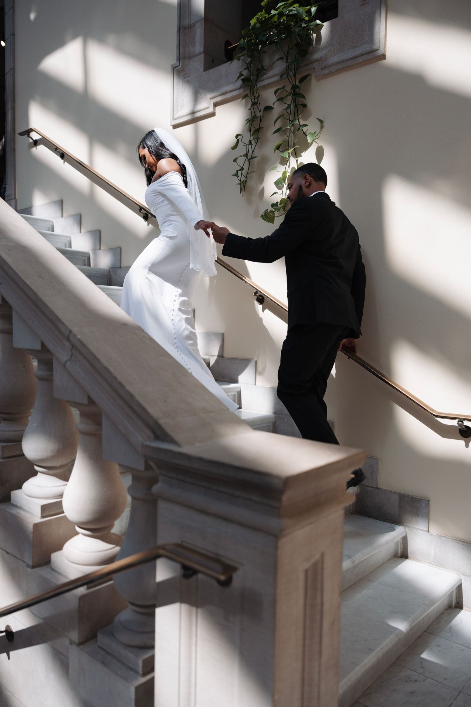 Bride and groom walking up elegant marble staircase in sunlit venue with greenery.