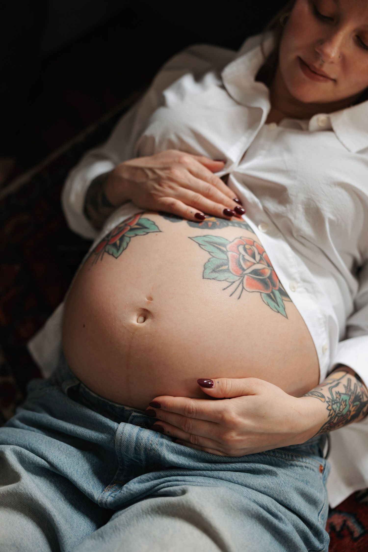Pregnant woman in white shirt and jeans cradling tattooed baby bump with rose tattoos, dark background.