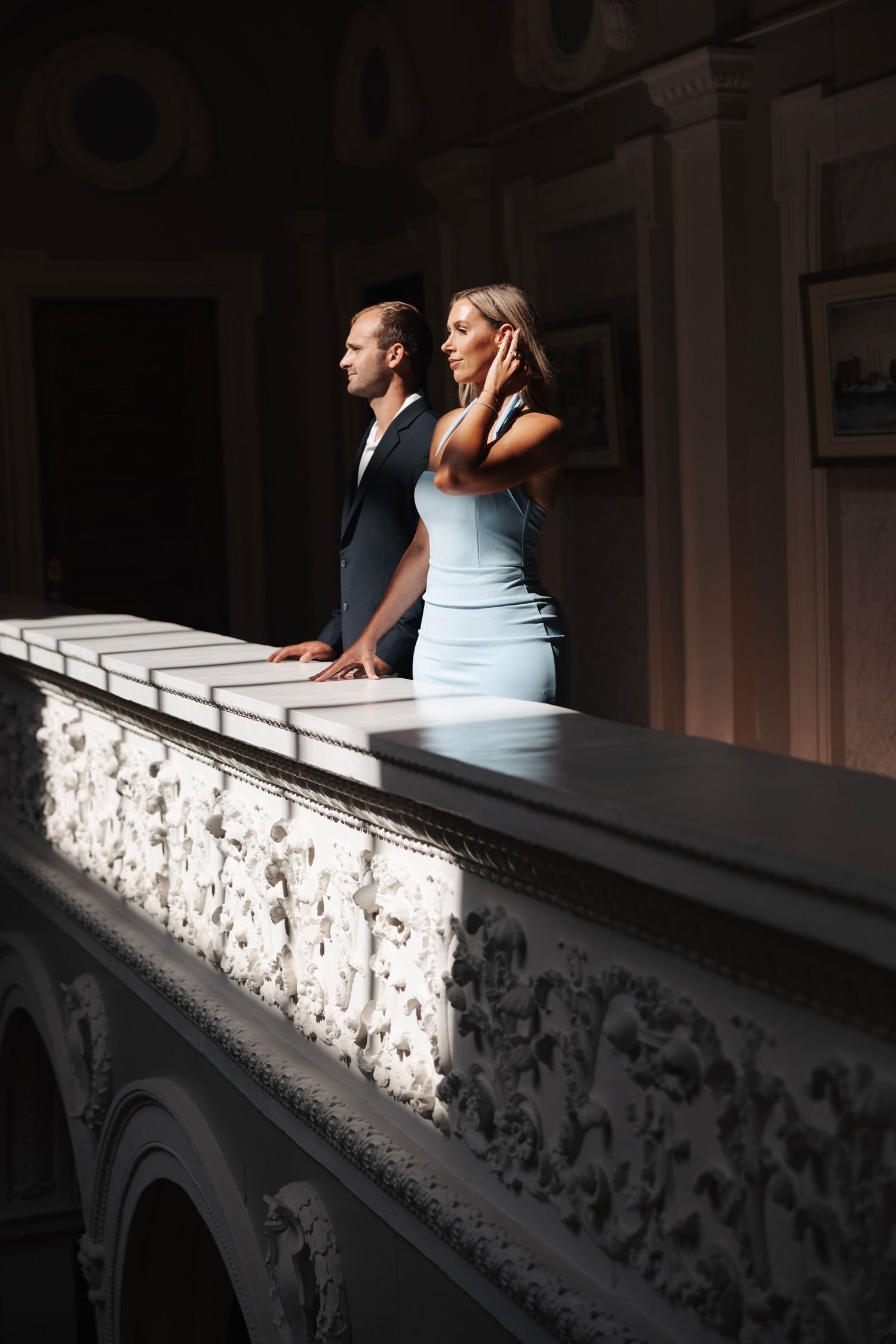 Elegant couple in formal attire standing at ornate balcony railing in dramatic interior lighting.