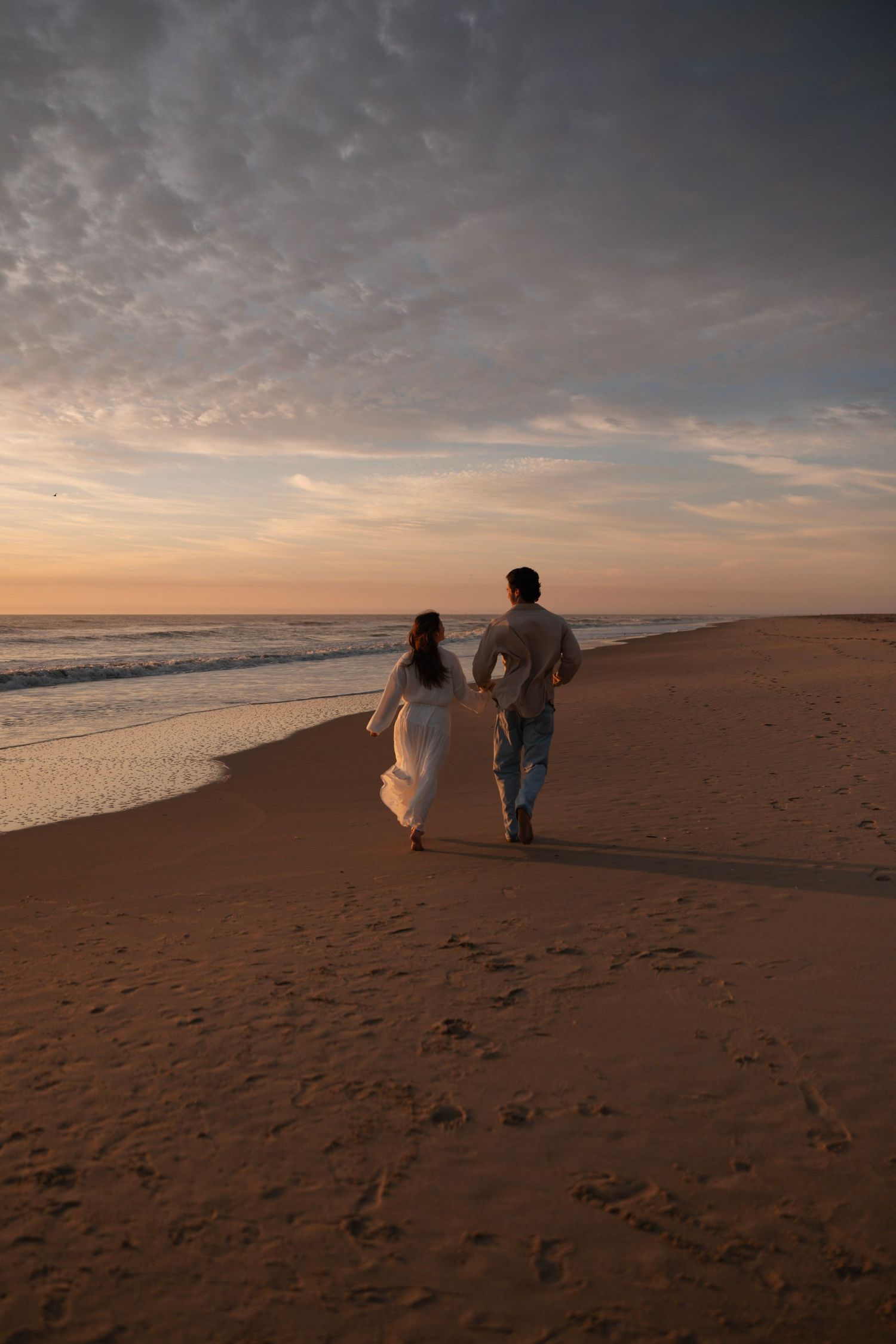 Couple in white dress and casual attire walking hand in hand on sandy beach at sunset.