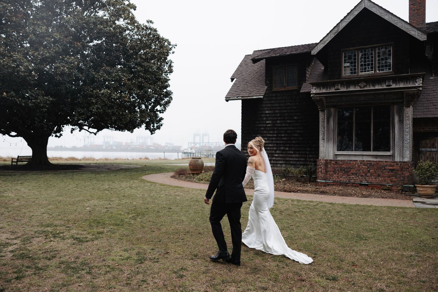 Bride and groom walk toward rustic dark shingled estate home surrounded by misty waterfront landscape.
