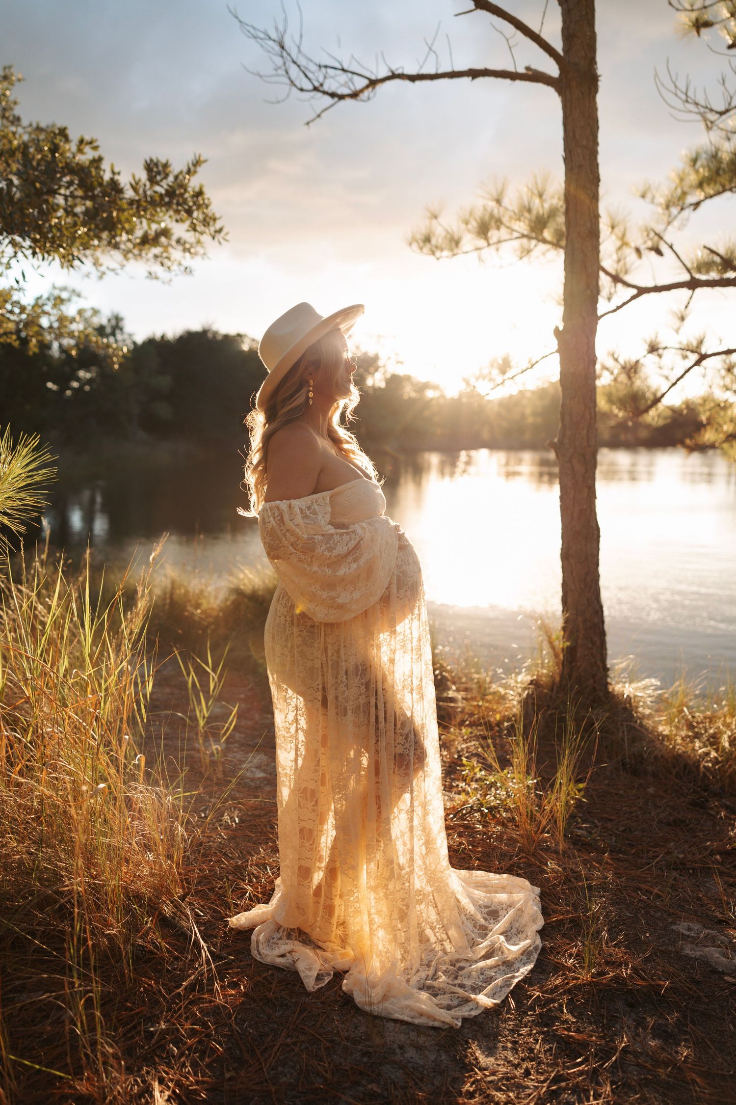 Pregnant woman in lace gown and hat standing by lake in golden sunset light.