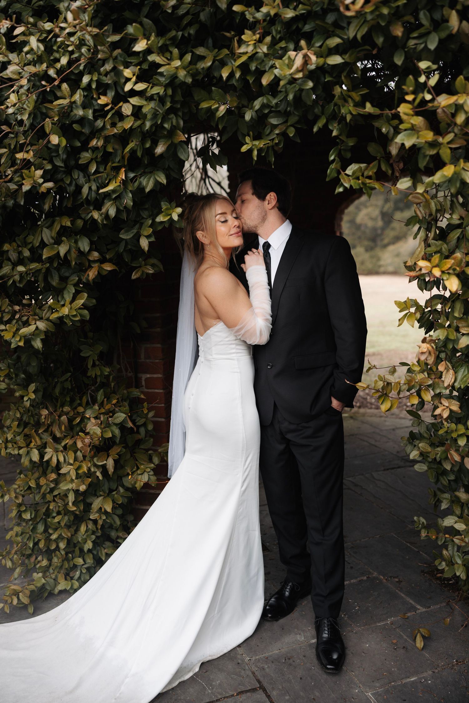 Bride in sleek white gown with veil and groom in black suit kissing under green leafy arch.