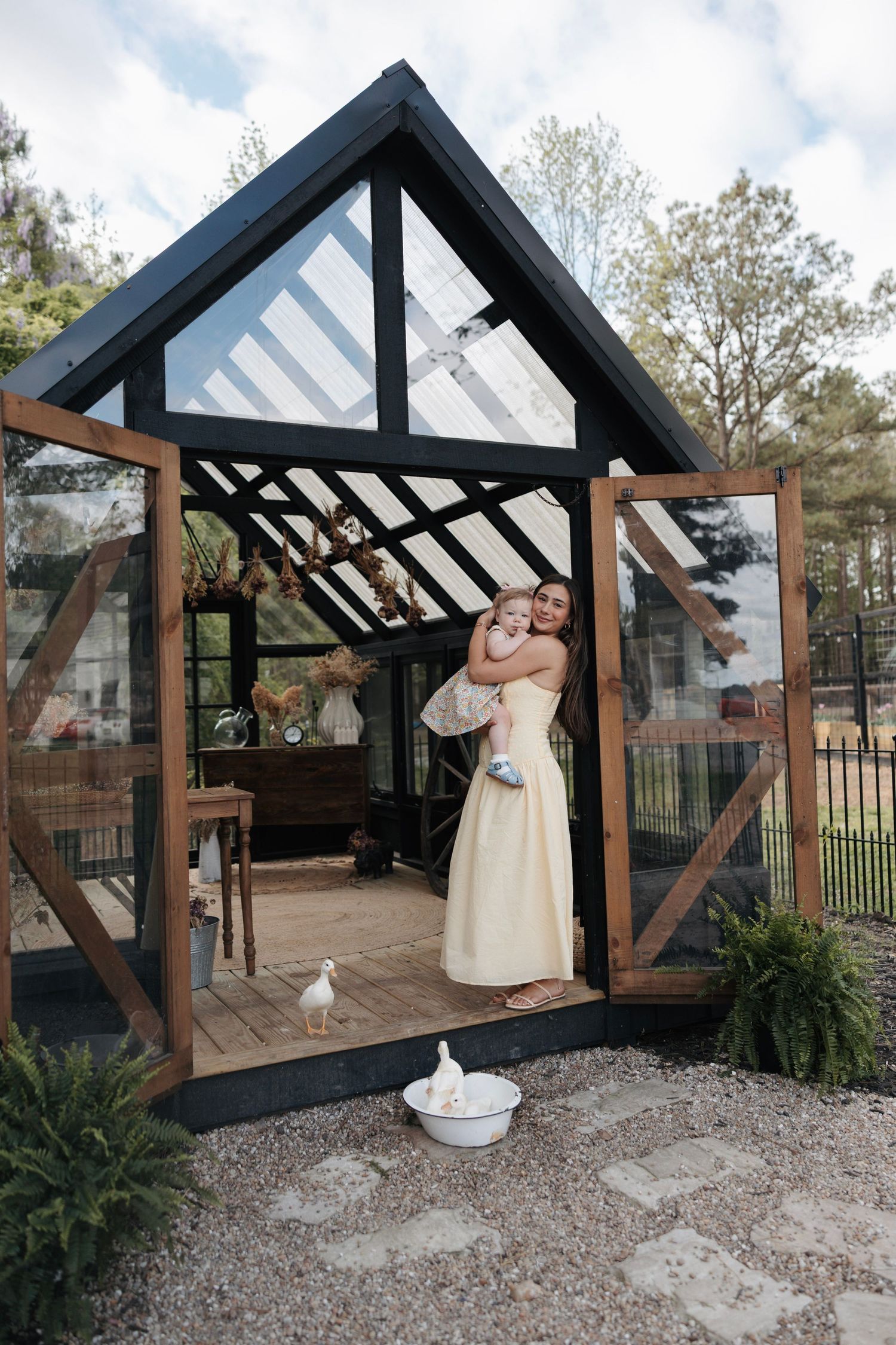 Couple embracing in doorway of black-framed glass greenhouse with white doves on gravel path.
