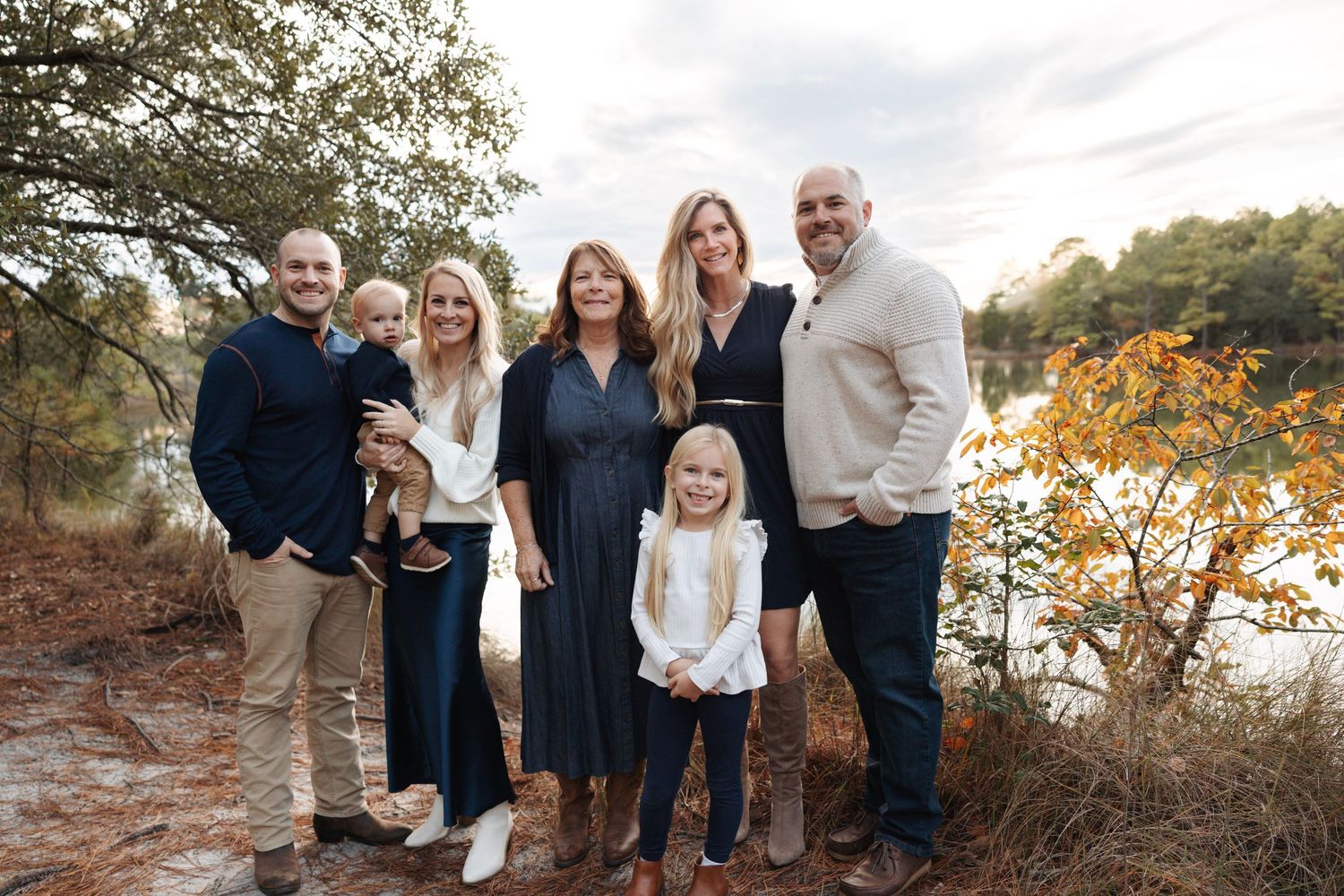 Happy extended family posing together outdoors near a lake surrounded by autumn foliage.