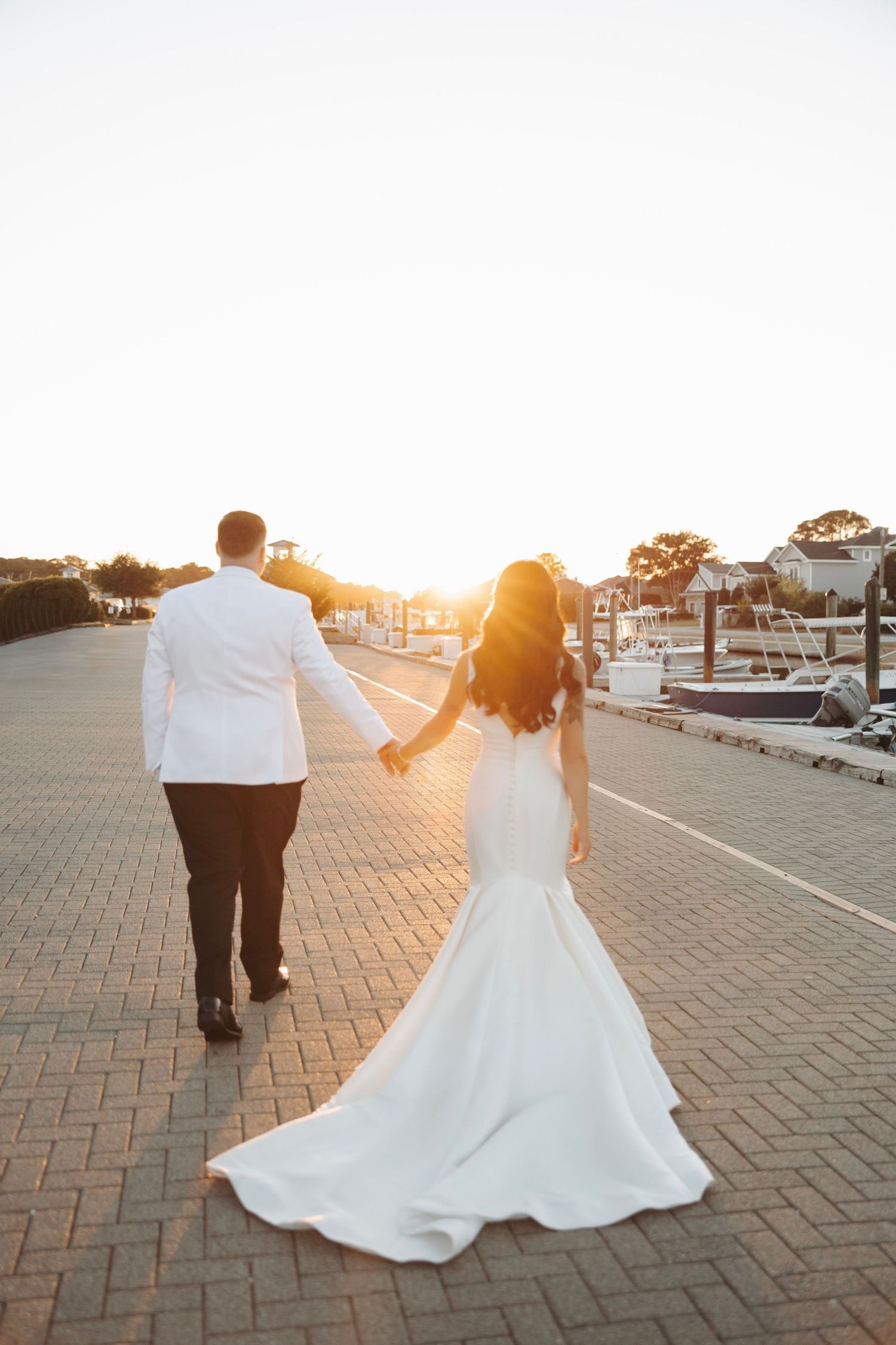 Bride and groom walk hand in hand along marina boardwalk at golden sunset, bride in flowing white gown.