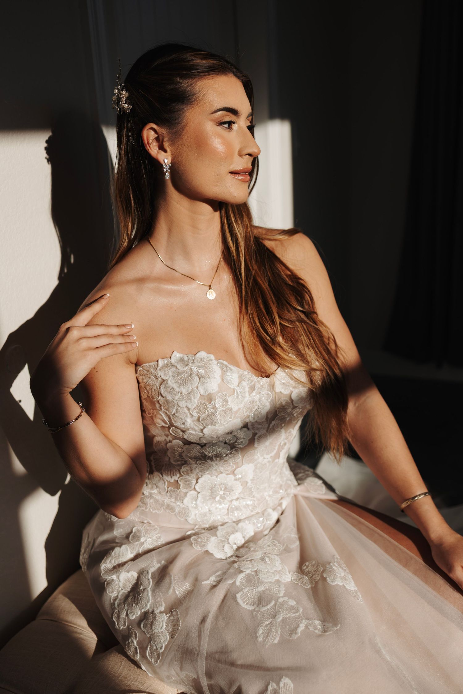 Elegant bride in strapless floral lace gown poses in dramatic sunlight with pearl jewelry.