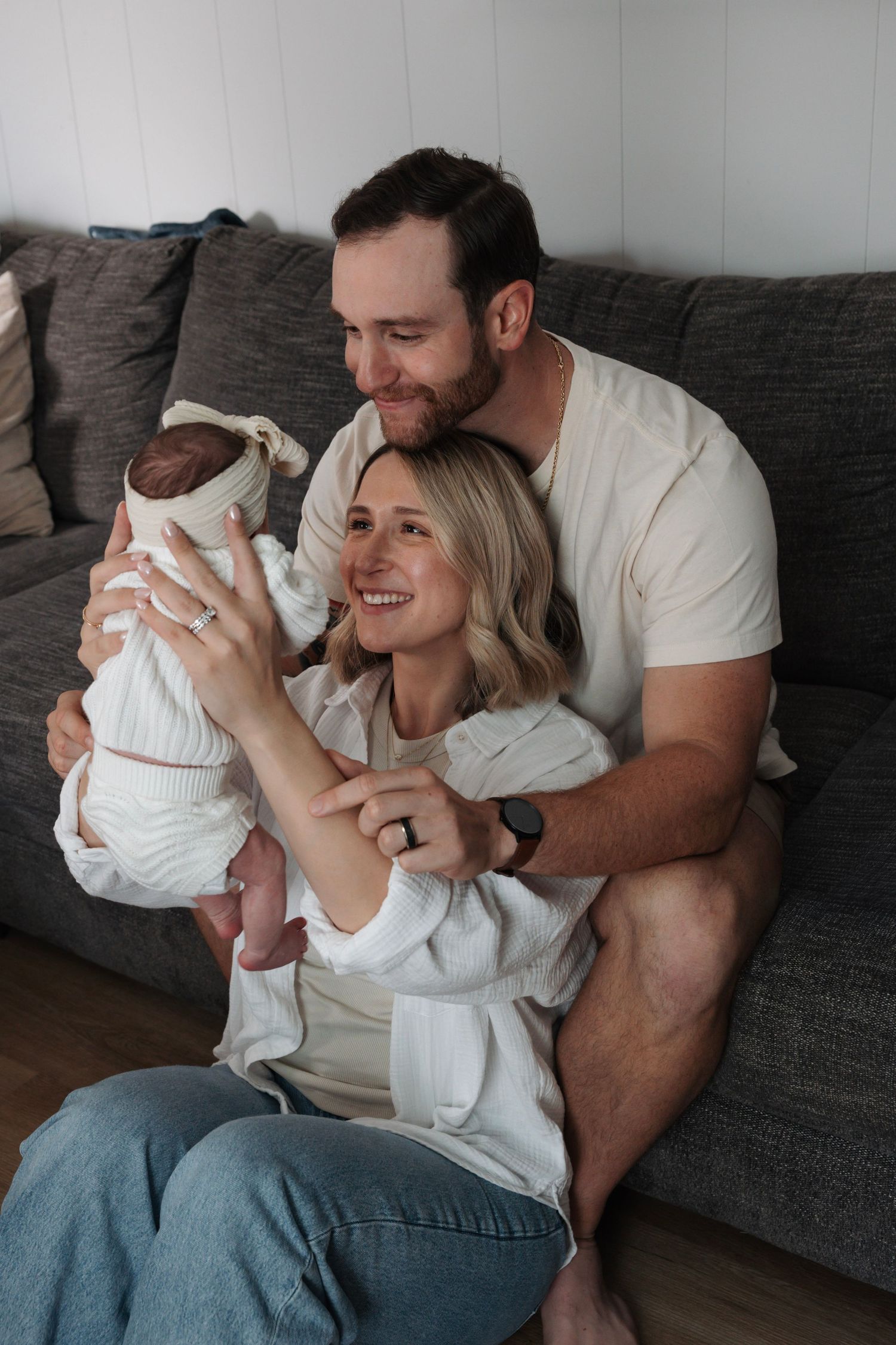 Joyful new parents sitting on gray sofa, smiling while holding their swaddled newborn baby.