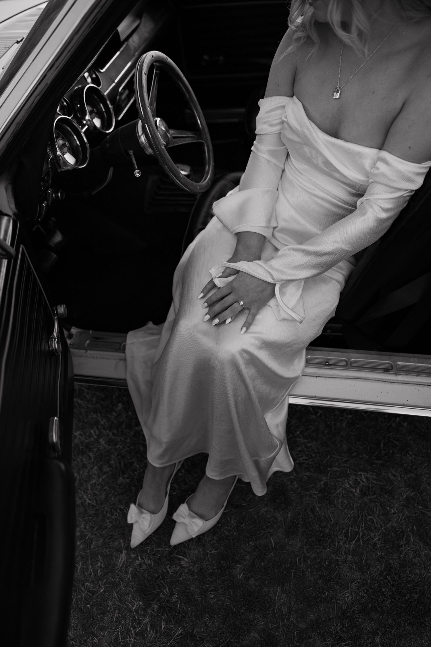 Black and white photo of bride in off-shoulder satin gown sitting in vintage car doorway.