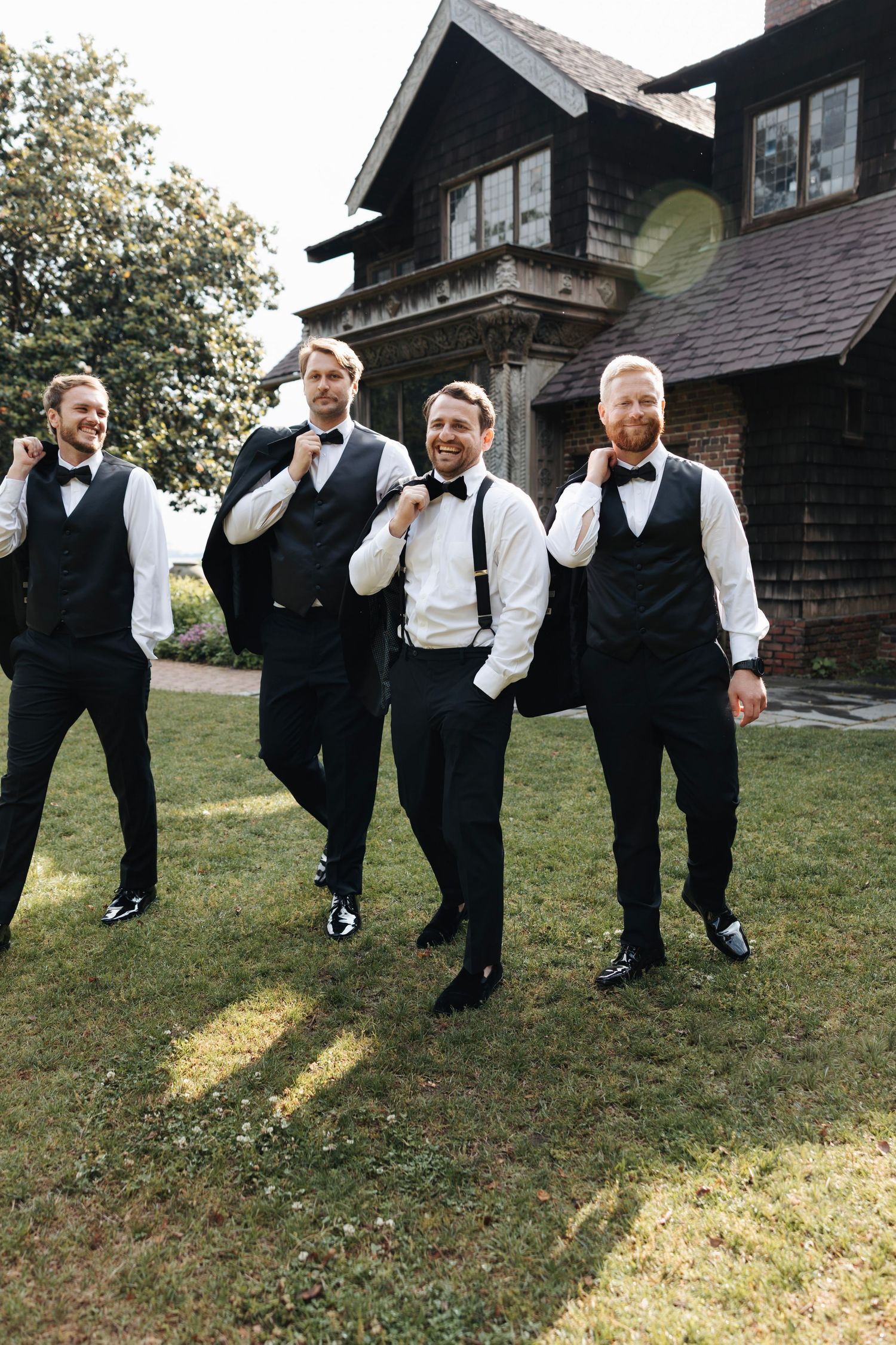 Four groomsmen in black vests and bow ties posing playfully outside a rustic house.