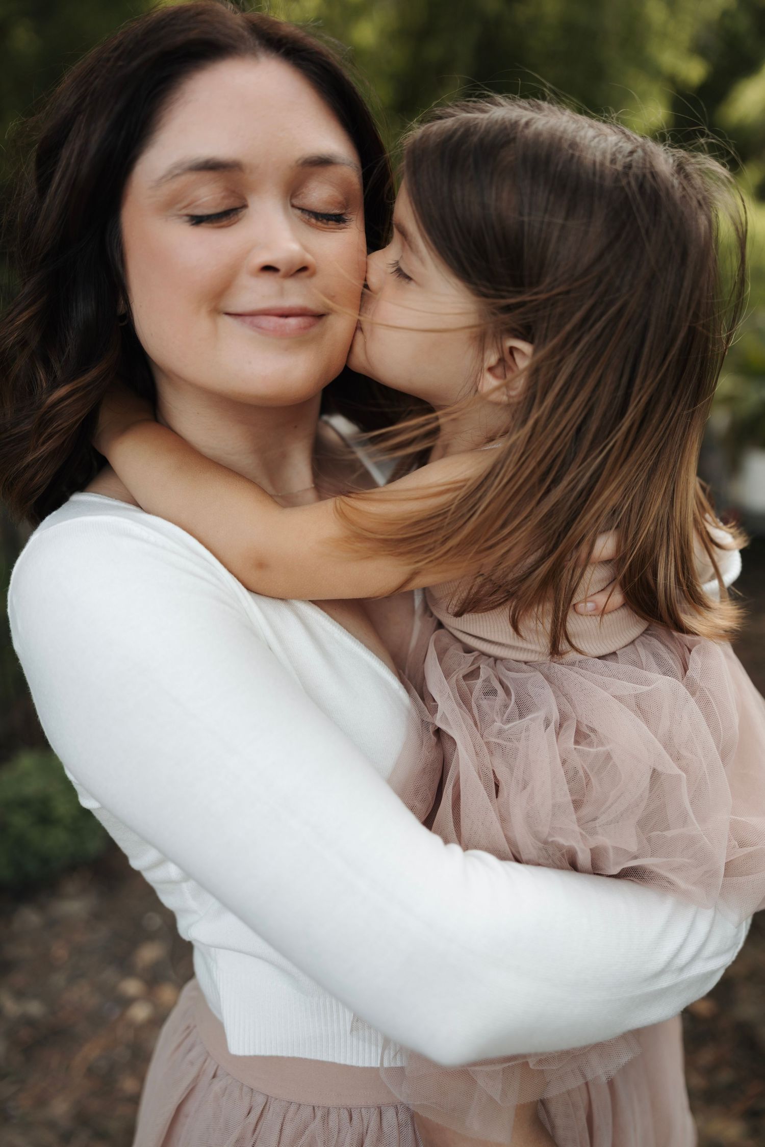 Young girl in pink tulle dress kissing smiling mother on cheek in outdoor garden setting.