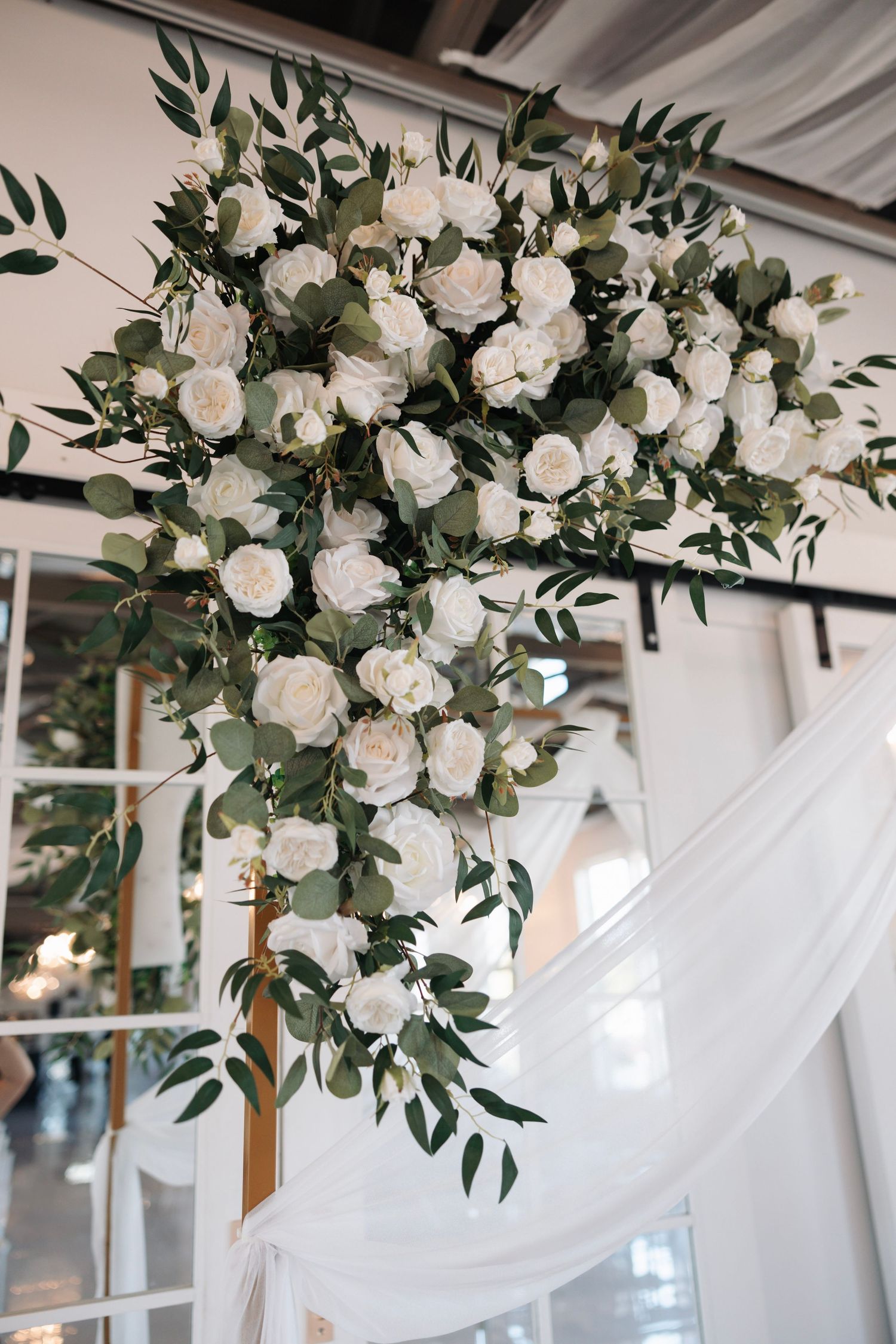 Lush wedding arch arrangement of white roses and eucalyptus greenery cascading from wooden frame indoors.