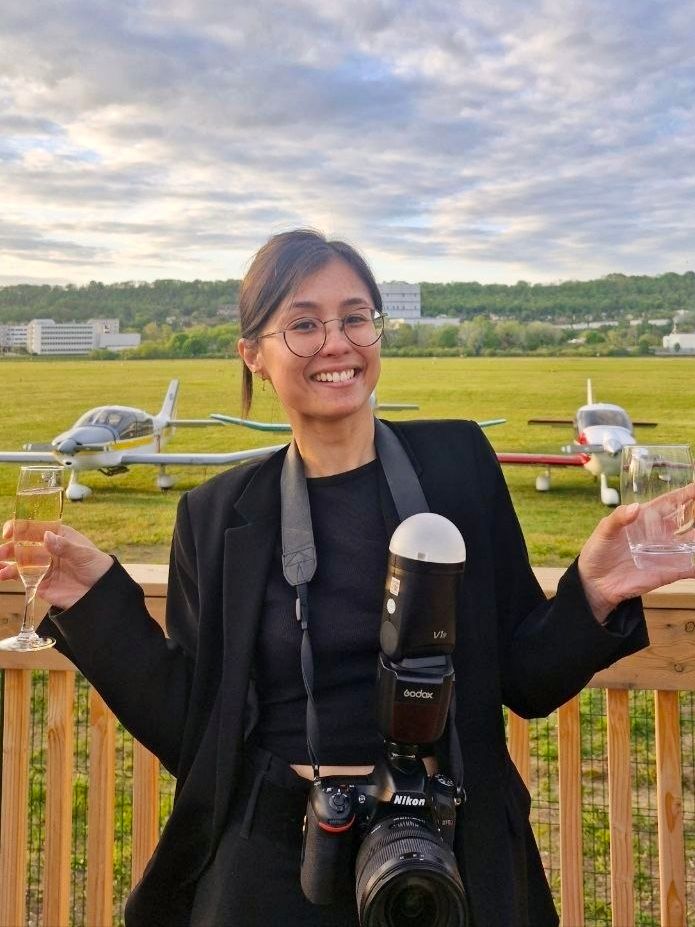 Femme souriante avec un appareil photo Nikon et un flash, debout sur le pont près d'un petit avion sur un aérodrome pendant l'heure dorée.