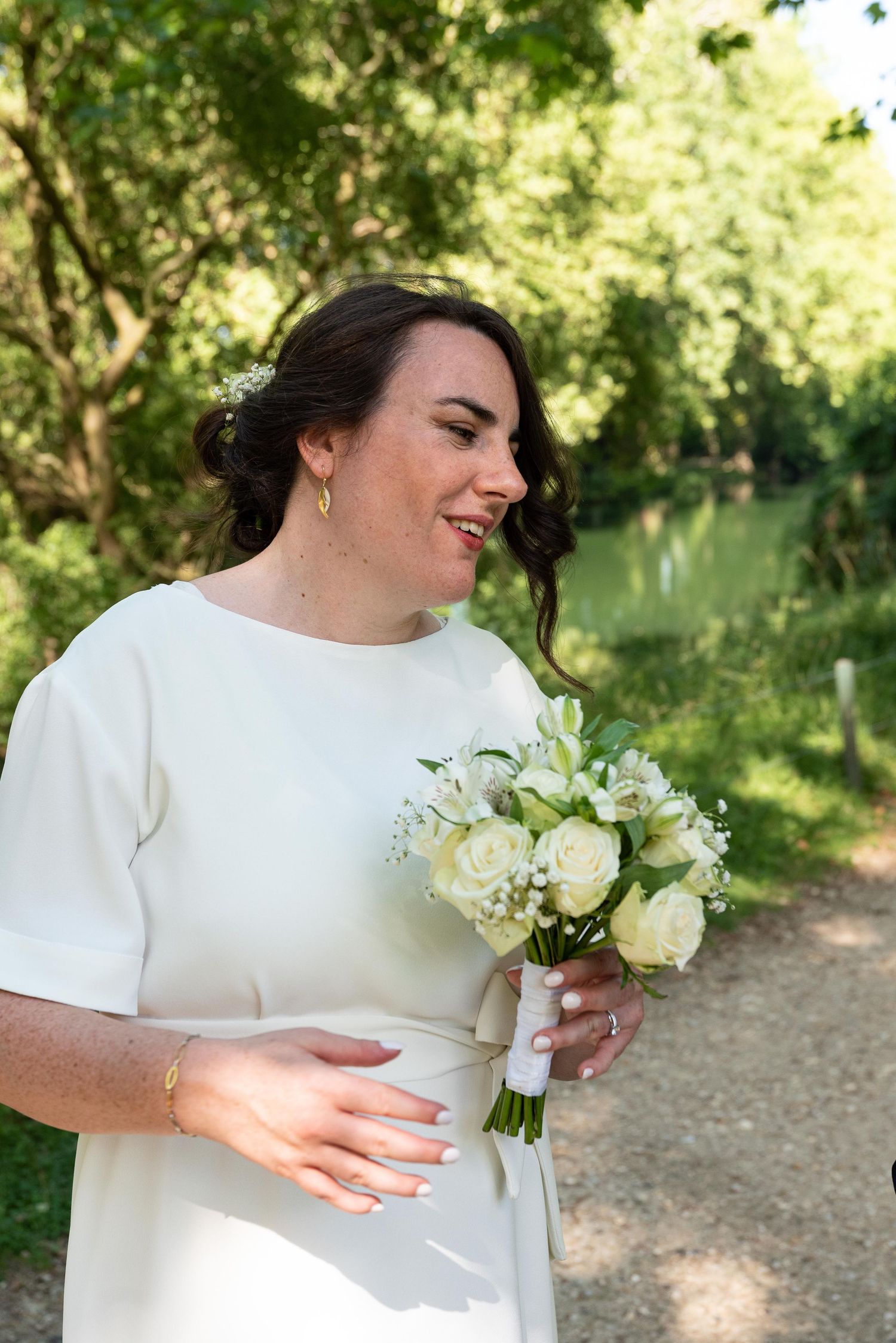 Une mariée radieuse en robe blanche, tenant un bouquet de roses blanches, sourit en plein air dans un parc verdoyant.