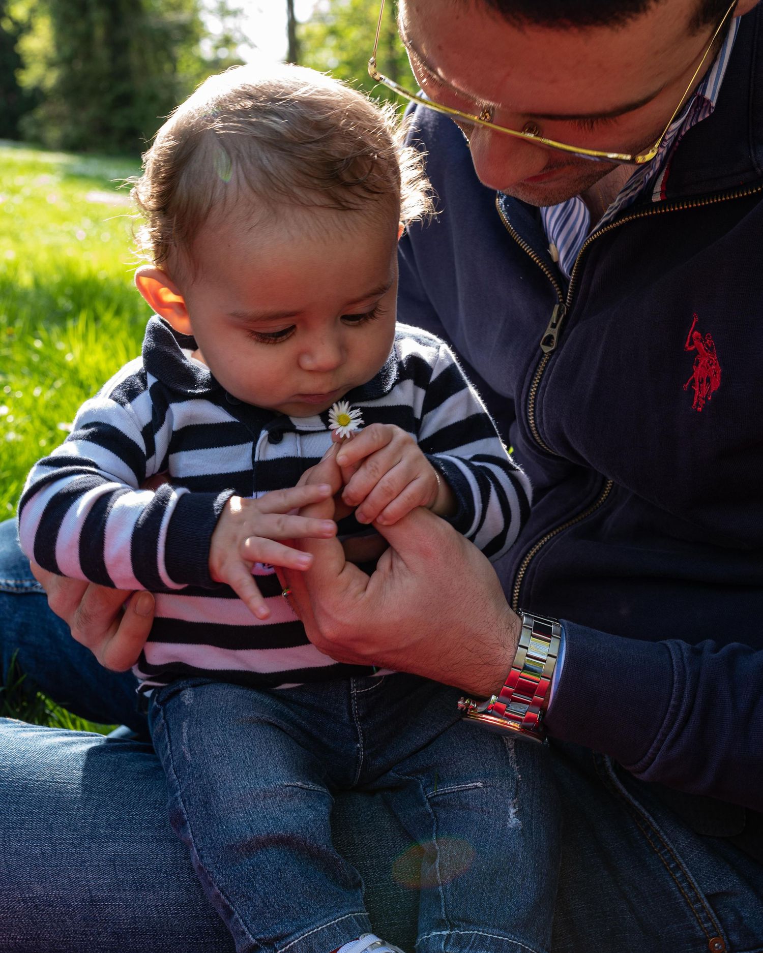 Father and baby sitting on grass examining a small white daisy flower together outdoors.