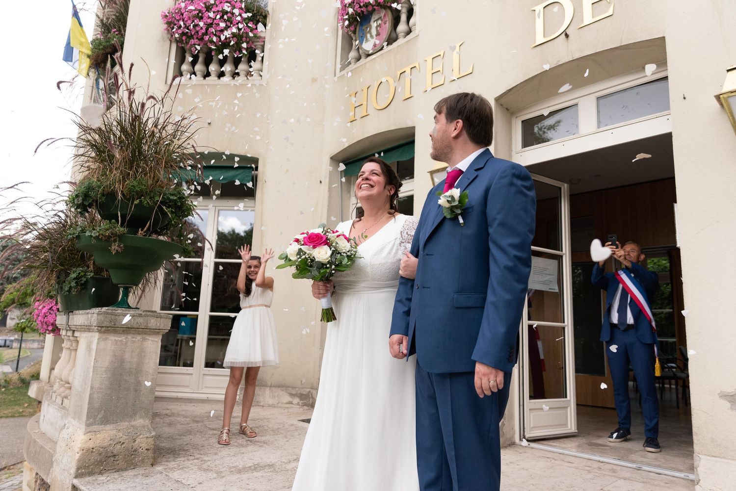 Un couple de jeunes mariés sourit devant l'Hôtel de Ville, la mariée tenant un bouquet rose et vêtue d'un tailleur bleu.