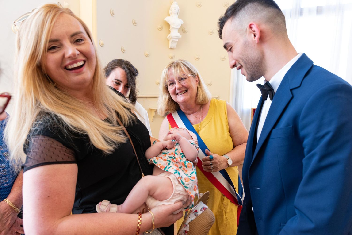 Une femme blonde souriante en robe noire tient un bébé lors d'une cérémonie formelle en intérieur, entourée d'invités.