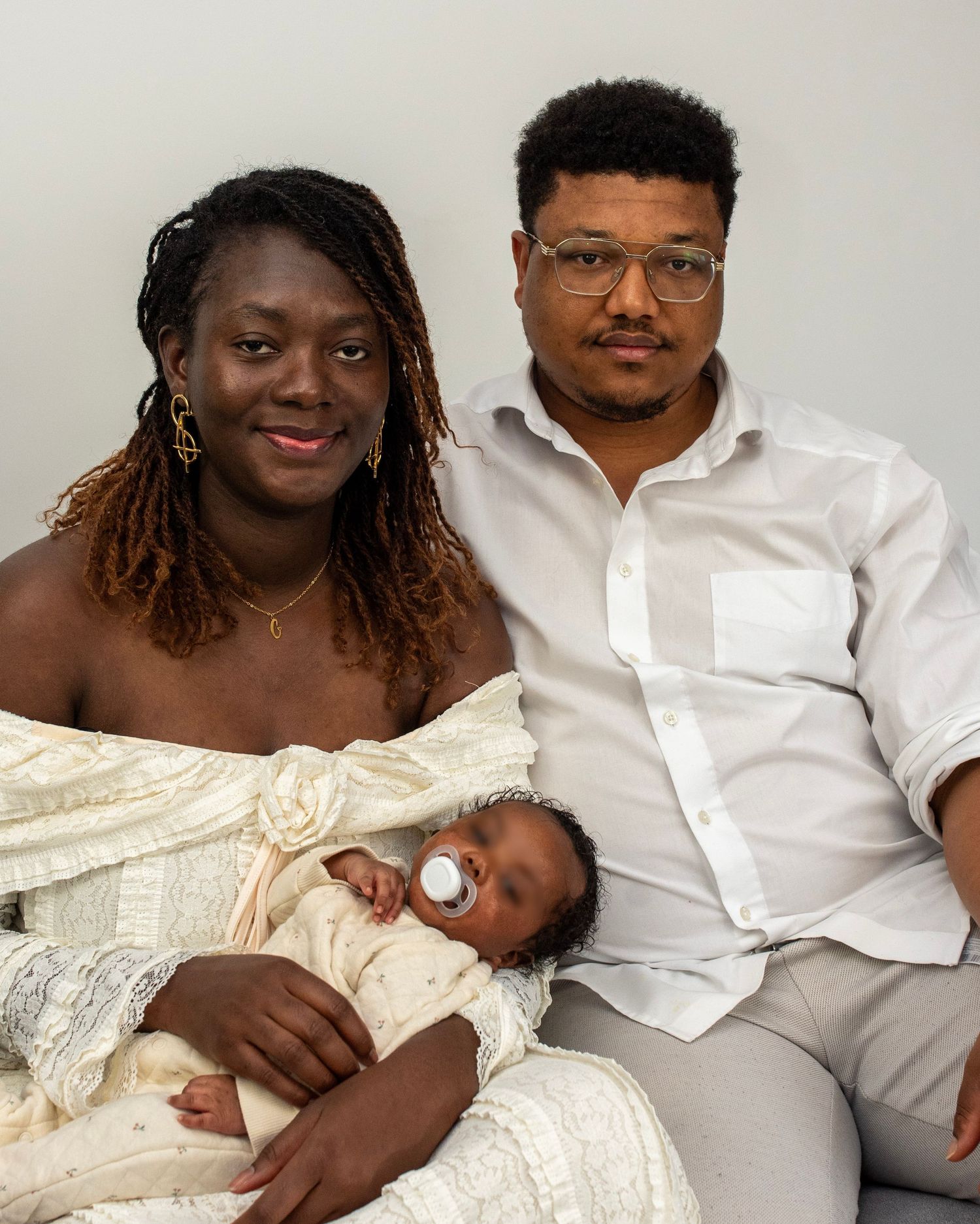 Un couple noir souriant pose avec leur nouveau-né sur un canapé dans une pièce d'un blanc éclatant.