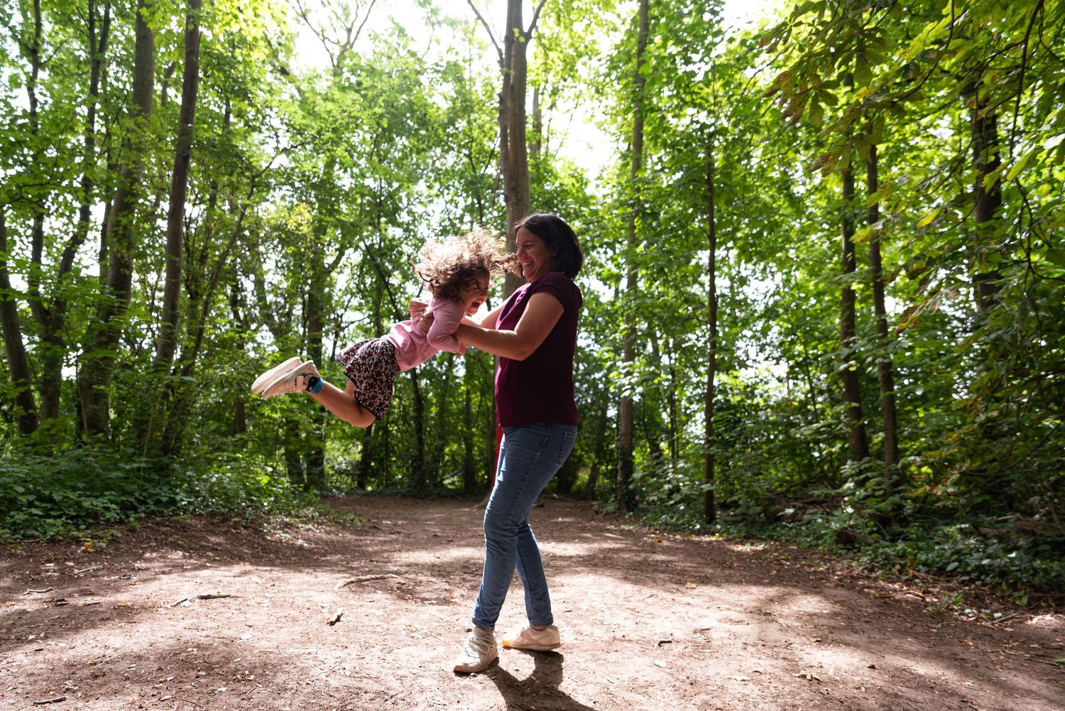 Une mère soulève et fait tournoyer son enfant sur un chemin forestier ensoleillé.