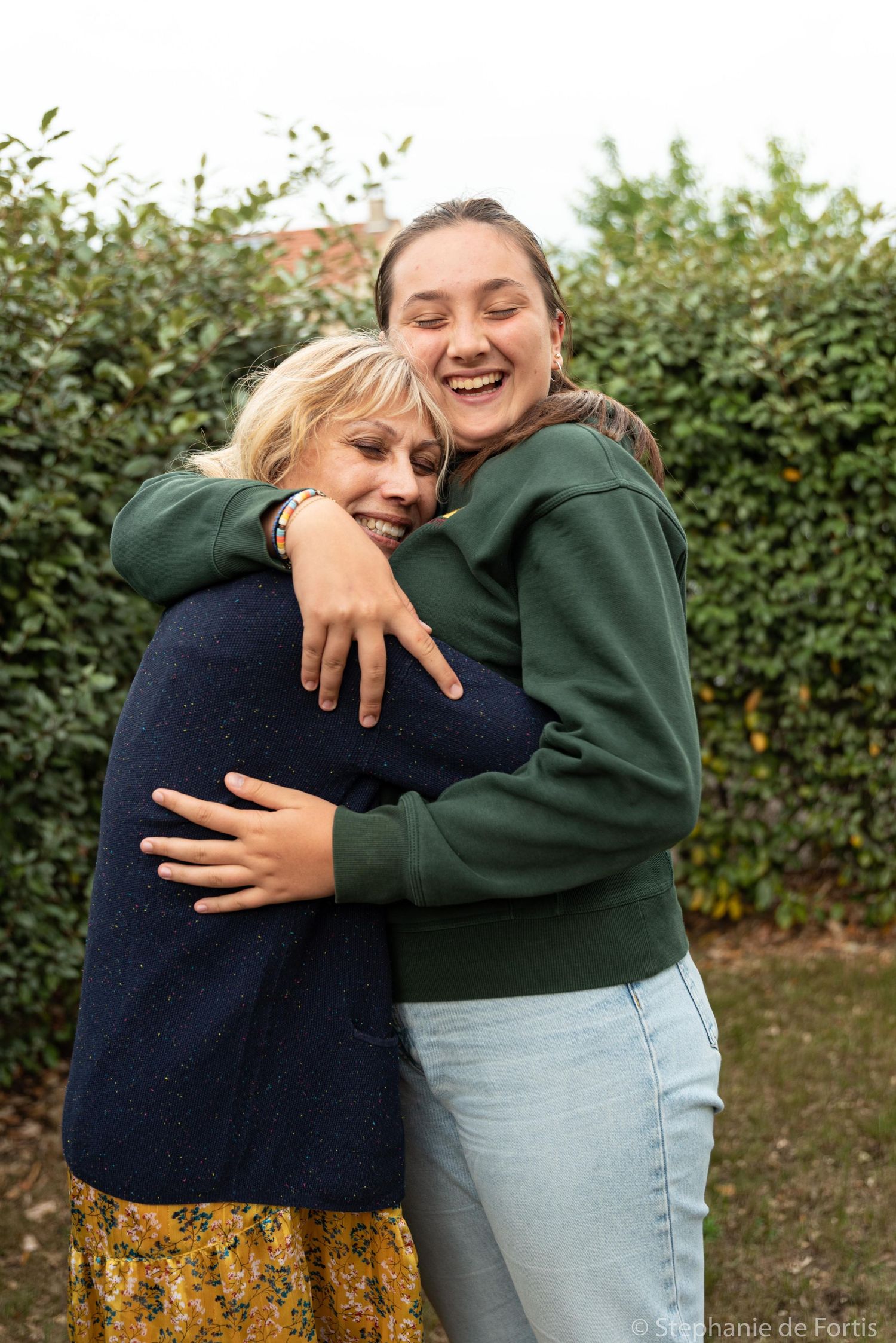 Deux femmes s'enlaçant chaleureusement en plein air, l'une riant joyeusement, entourées de haies vertes.
