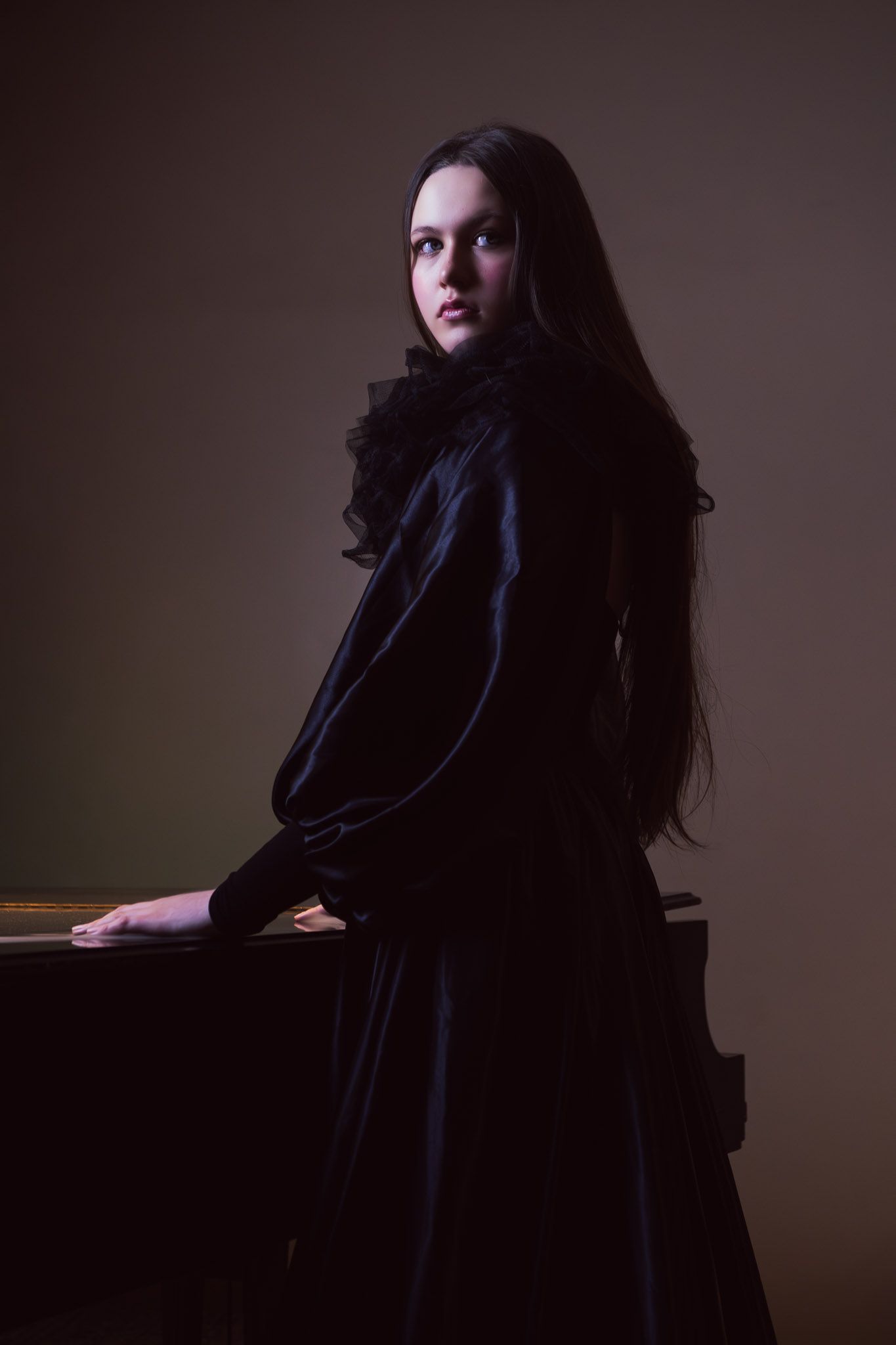 A young woman with long dark hair wearing an elegant black Victorian dress, posing dramatically in a dimly lit studio.