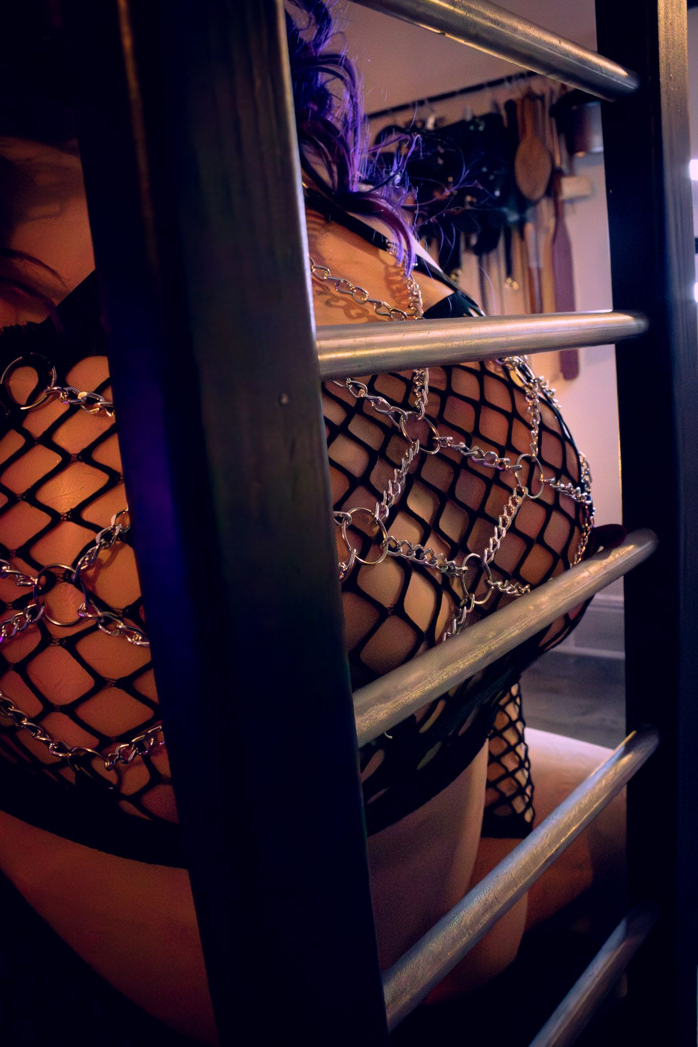 Person in fishnet bodysuit with chains poses behind metal bars in dramatic moody lighting.