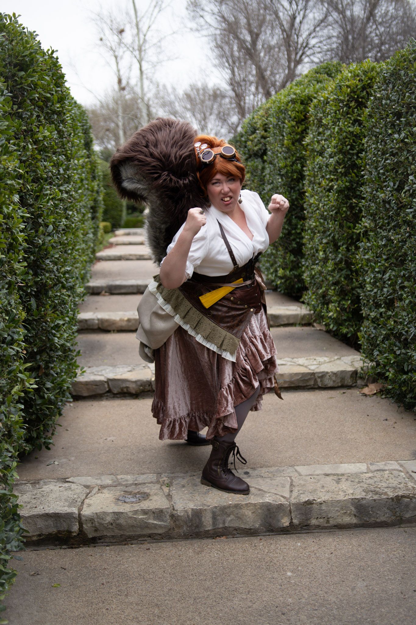 Woman in steampunk Squirrel Girl costume with goggles and ruffled skirt poses triumphantly on garden pathway lined with hedges.