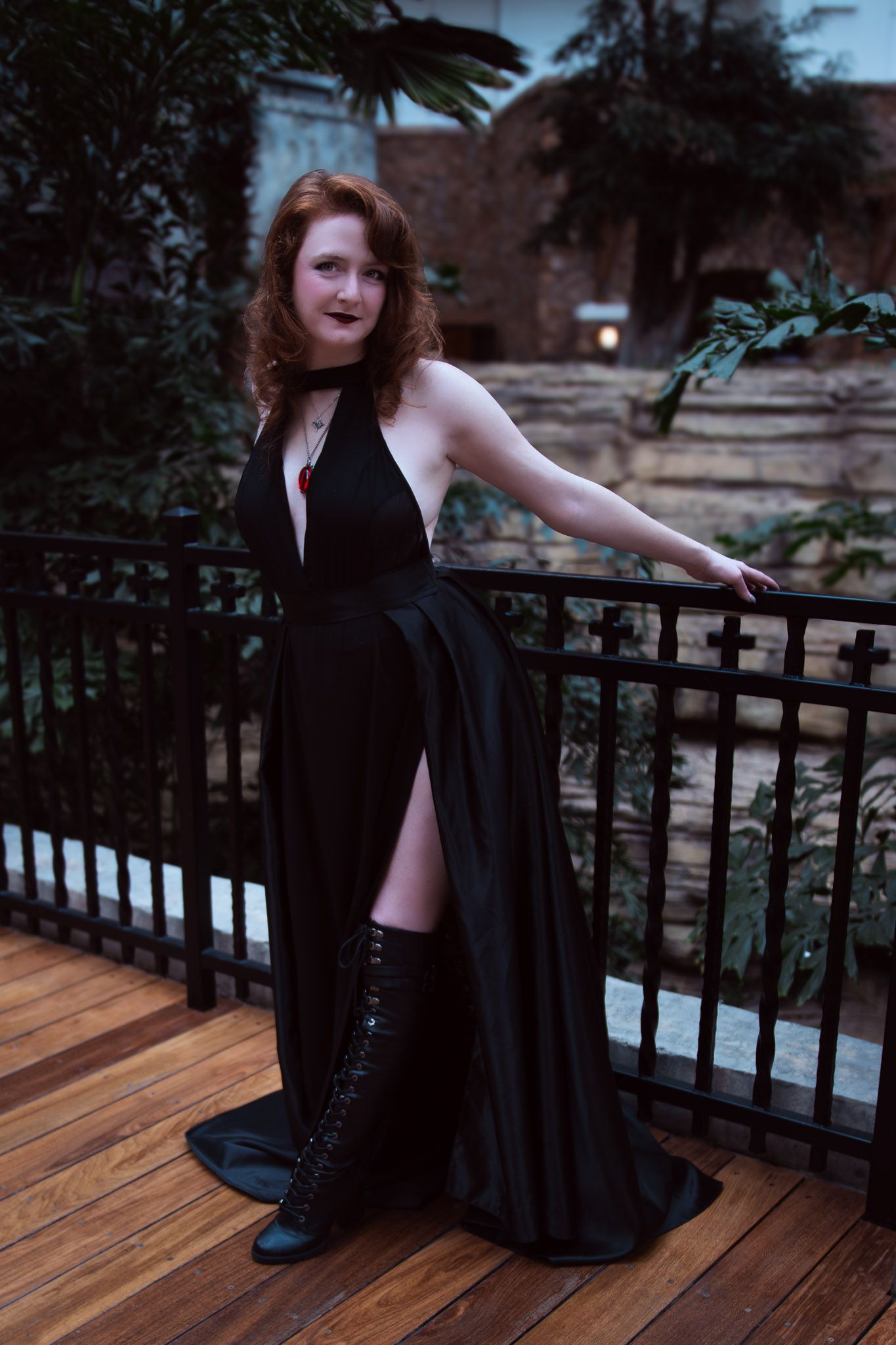 Woman in black halter gown with thigh slit and lace boots poses on wooden deck balcony at dusk.