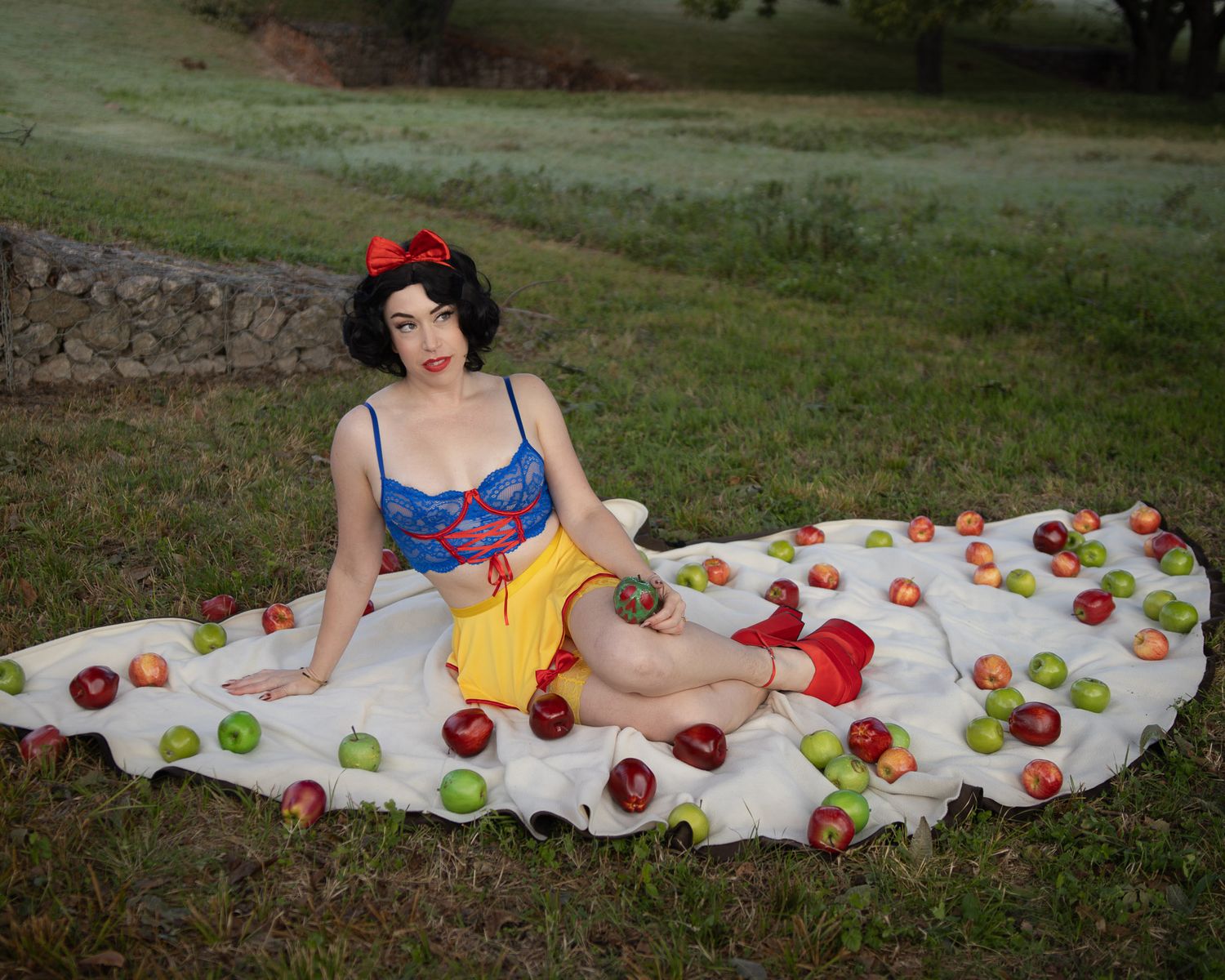 Woman in a Snow White costume with a red bow sits on a white blanket covered in red and green apples on a grassy field.