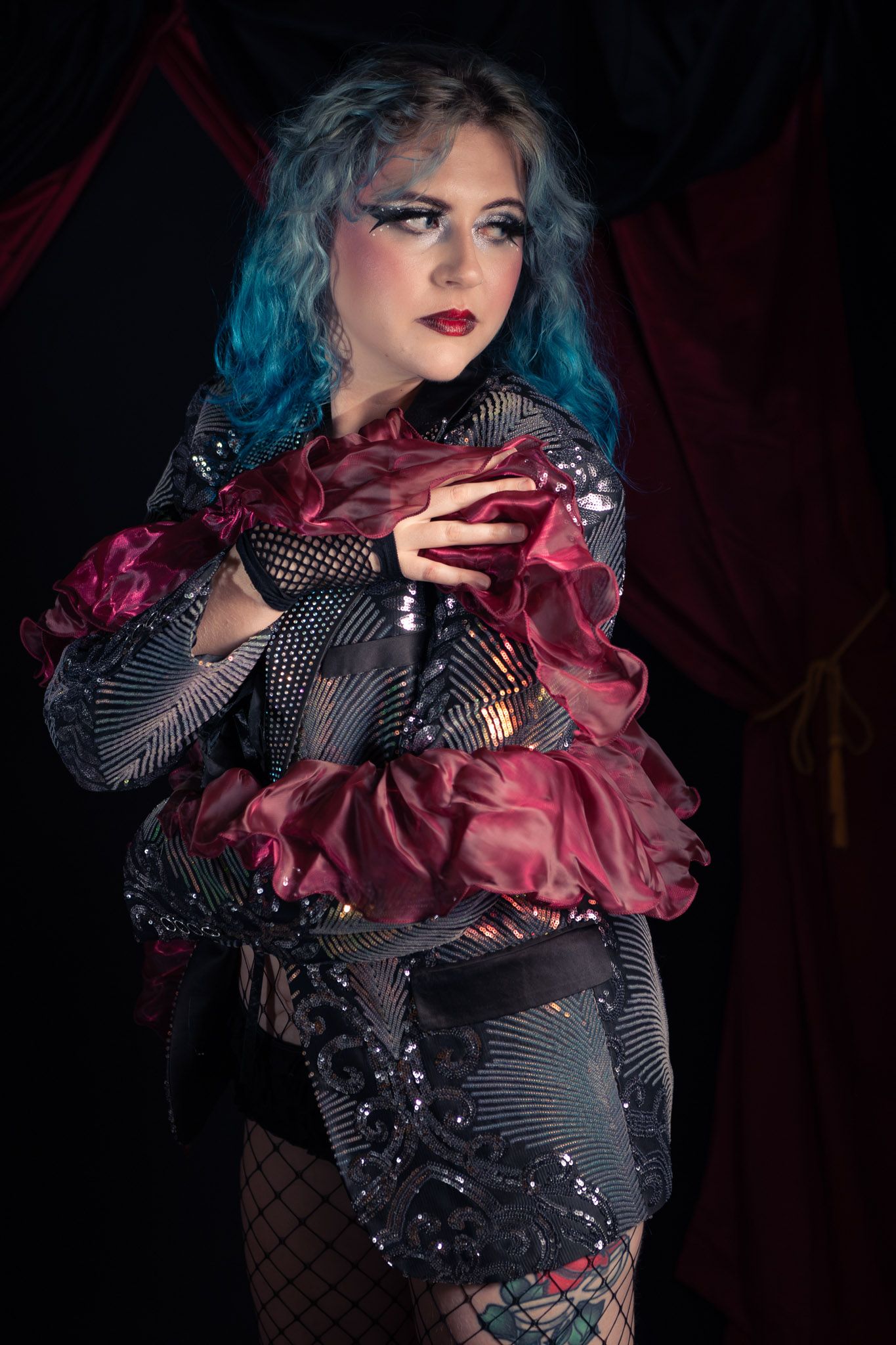 Woman with blue hair in dramatic burgundy and black sequined costume poses against dark red curtain backdrop.