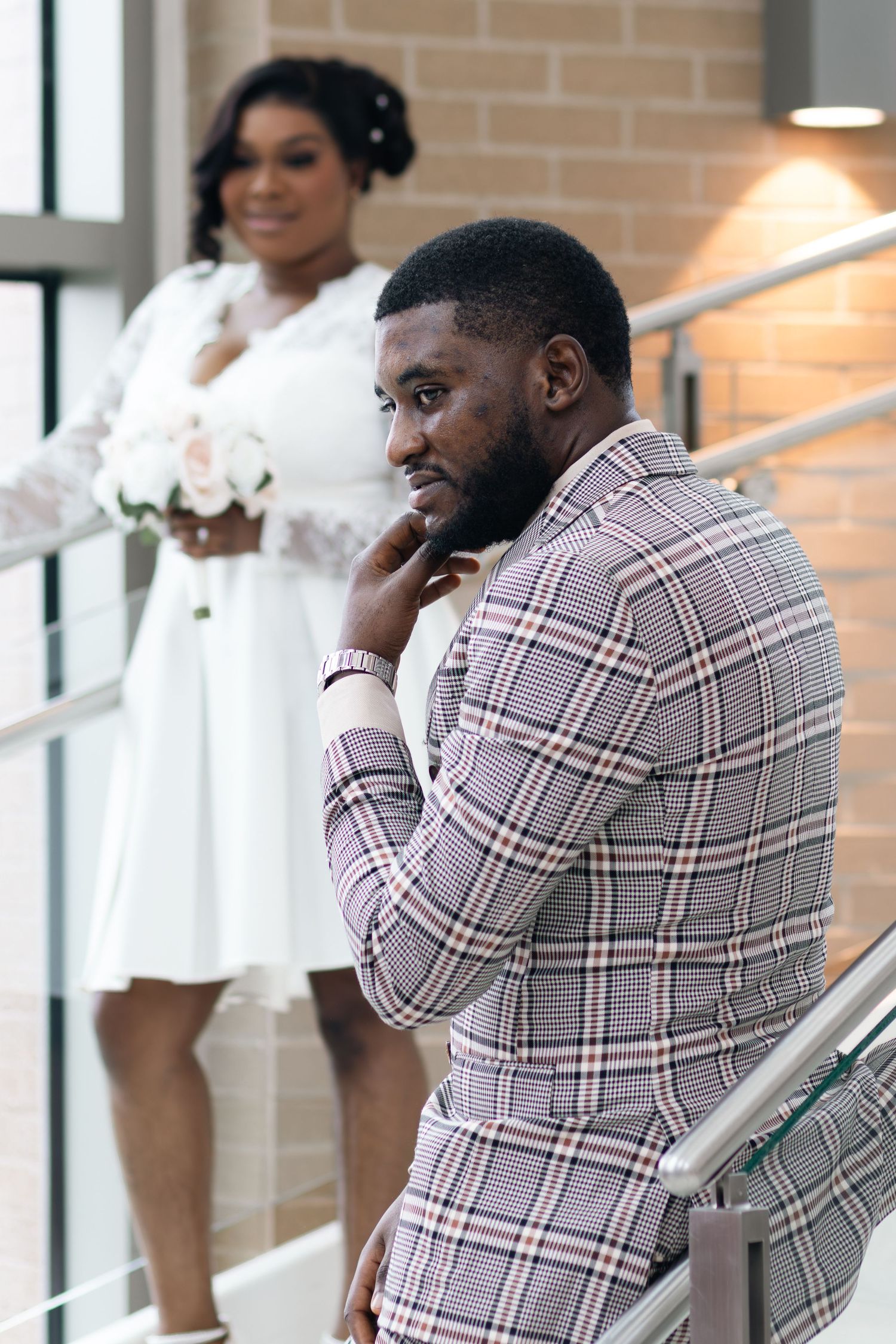 A groom in a plaid suit poses on stairs while his bride in a white dress holds a bouquet in the background.