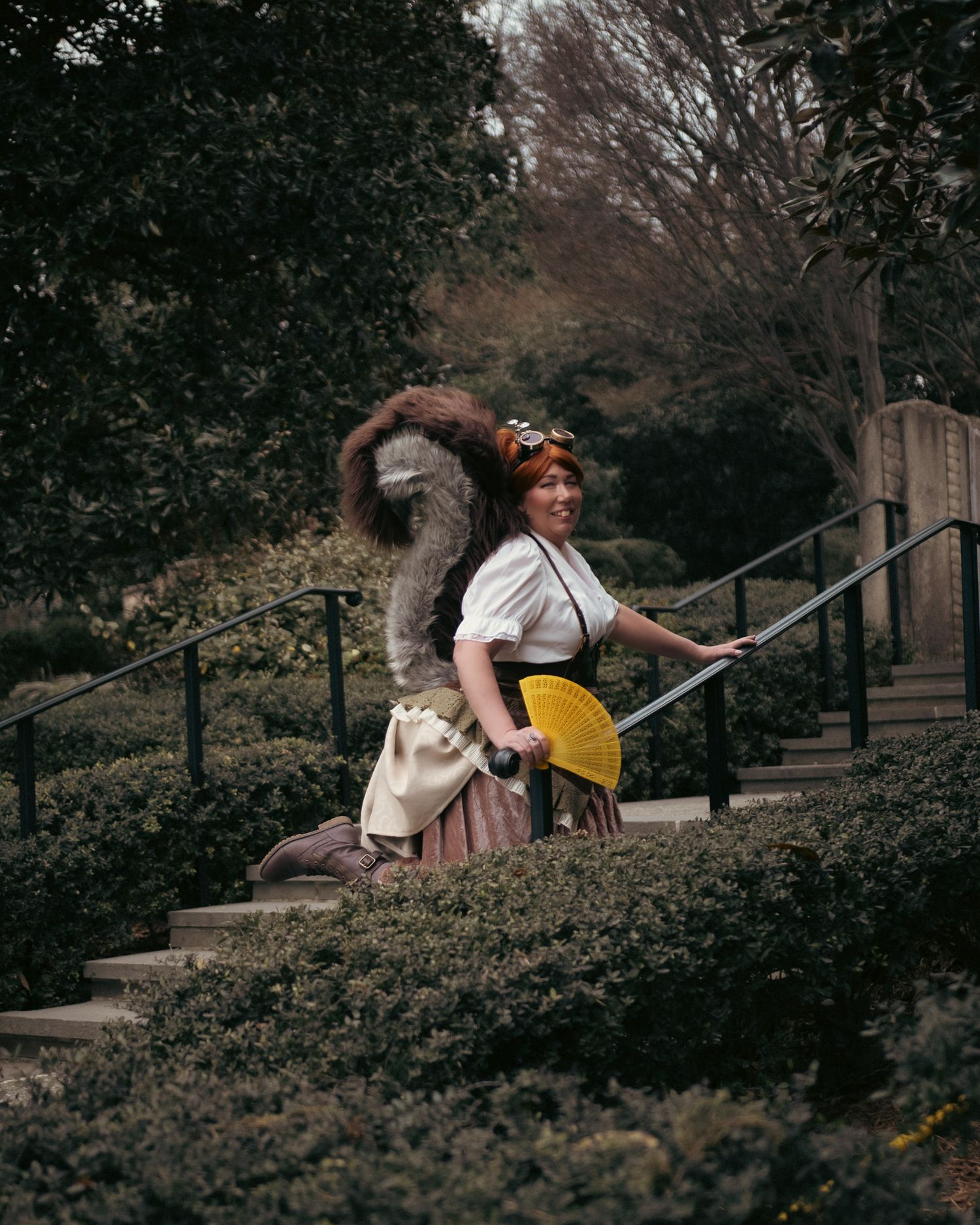 Woman in vintage costume with yellow fan and squirrel on stairs in garden.