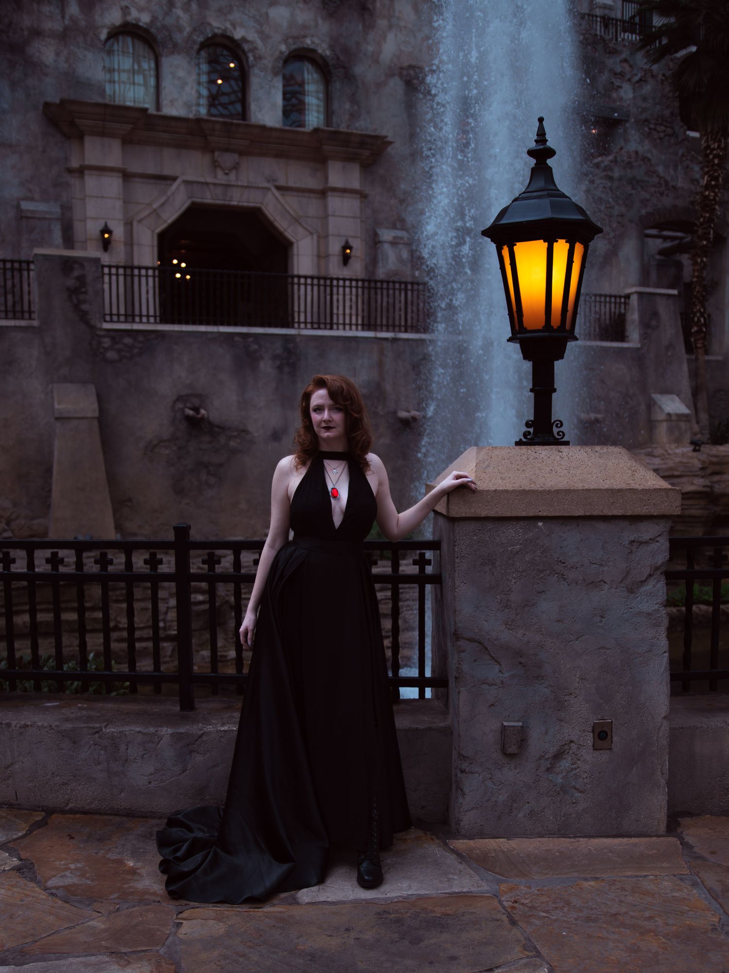 Woman in elegant black gown poses by glowing lantern with fountain and stone building backdrop at dusk.